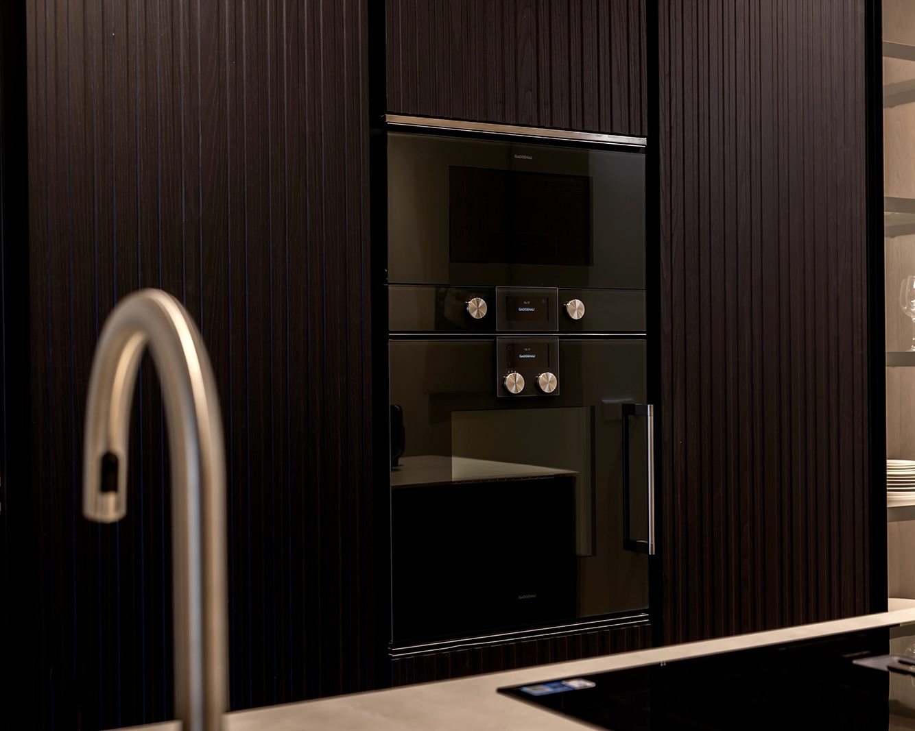 Cocina moderna con gabinetes oscuros y electrodomésticos empotrados. Se ve un horno y microondas integrados en el mueble, y parte de un grifo de cocina en primer plano. El diseño es minimalista y elegante.