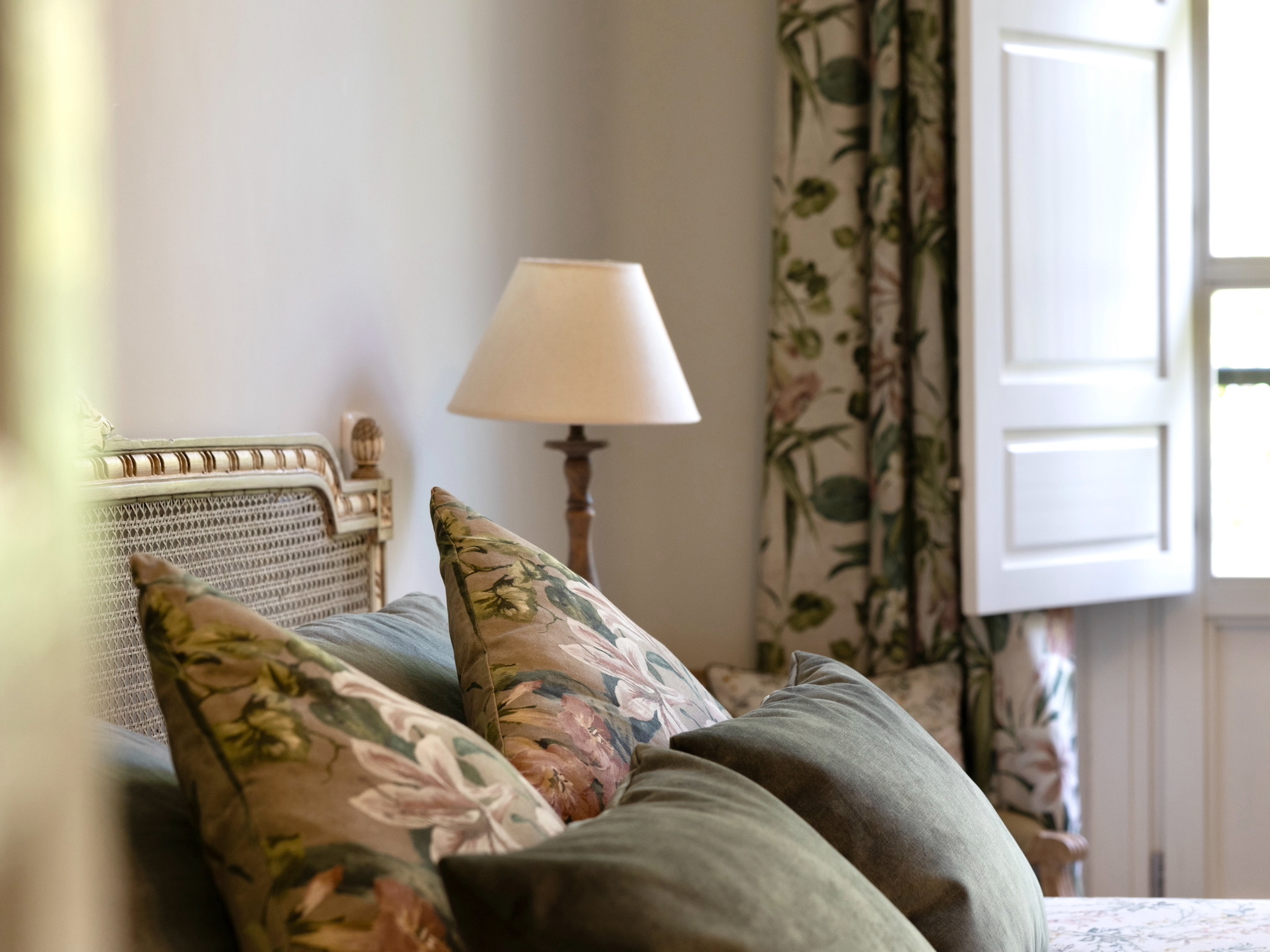 Dormitorio con decoración elegante estilo provenzal, cabecero de mimbre, cojines verdes y florales, lámpara blanca y cortinas con estampado botánico. La luz natural entra por la ventana creando una atmósfera acogedora.