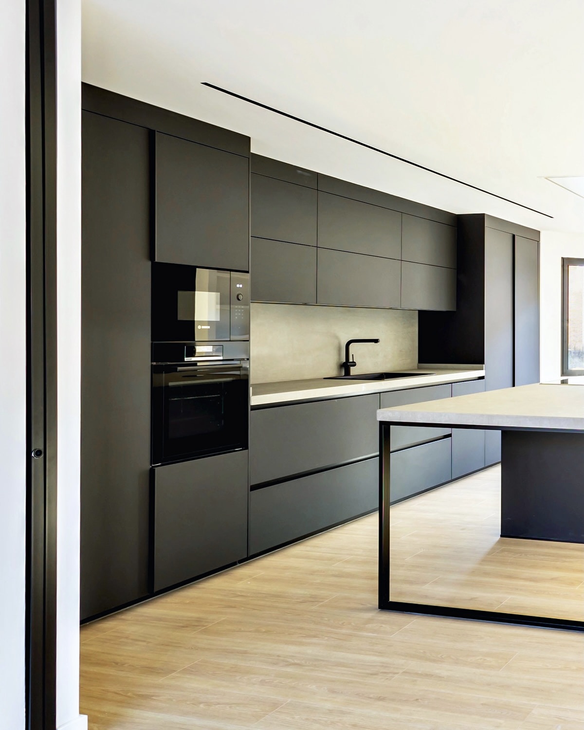 Cocina moderna y minimalista con gabinetes negros, encimera blanca, electrodomésticos integrados y una isla. El diseño es elegante y limpio, con líneas rectas y acabados mate.