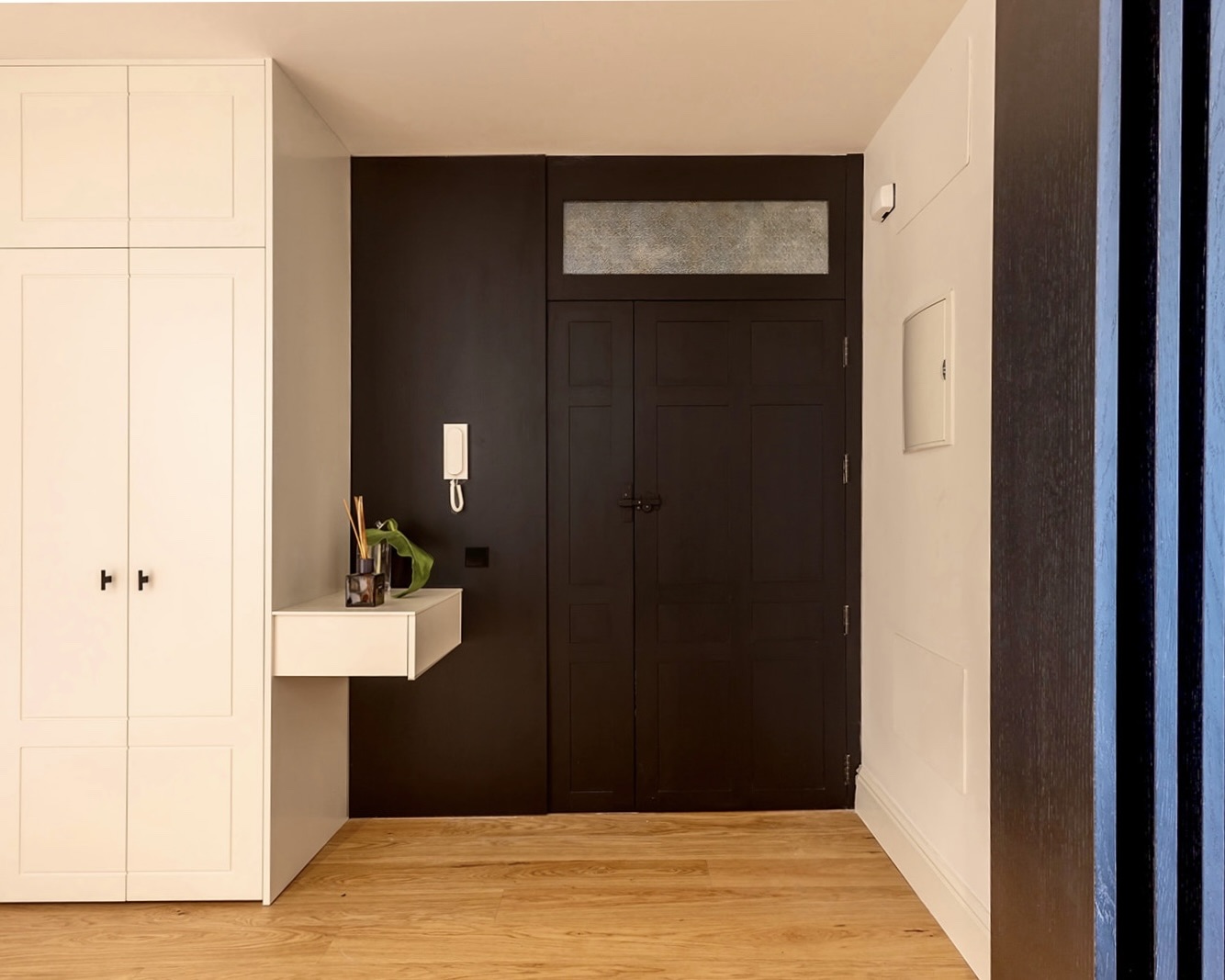 Entrada moderna de apartamento con puerta negra, armario blanco y suelo de madera. Contraste elegante entre blanco y negro. Pequeña consola blanca con objetos decorativos y comunicador en la pared.