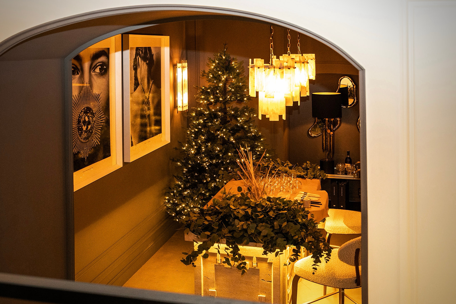 Vista a través de un arco de una sala decorada para Navidad con un árbol iluminado, lámparas colgantes, cuadros artísticos y una mesa con arreglos florales, creando una atmósfera elegante y festiva