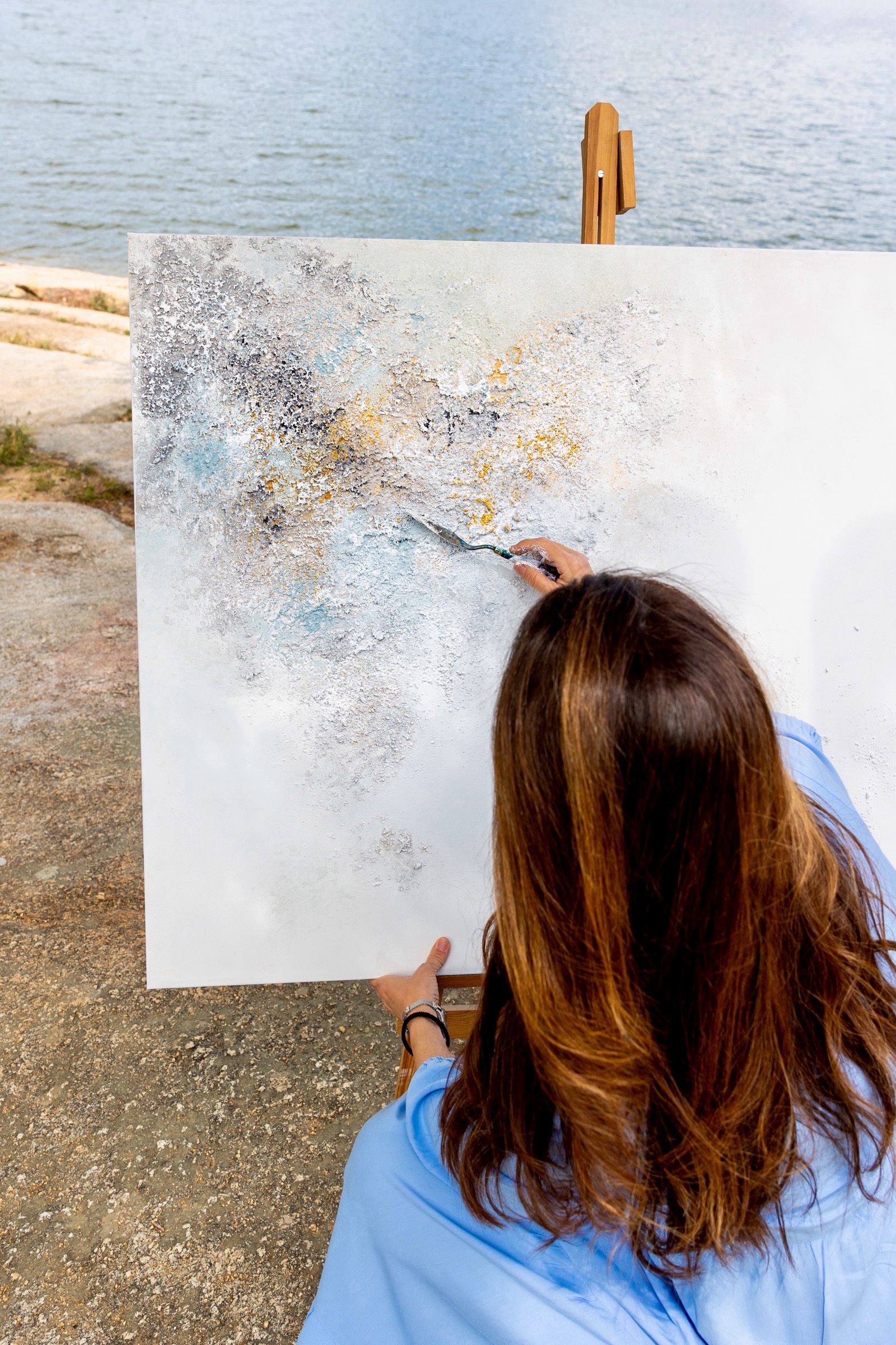 Artista pintando un cuadro abstracto en tonos azules, grises y dorados en un caballete junto al mar. La pintura refleja la textura y colores del agua cercana.