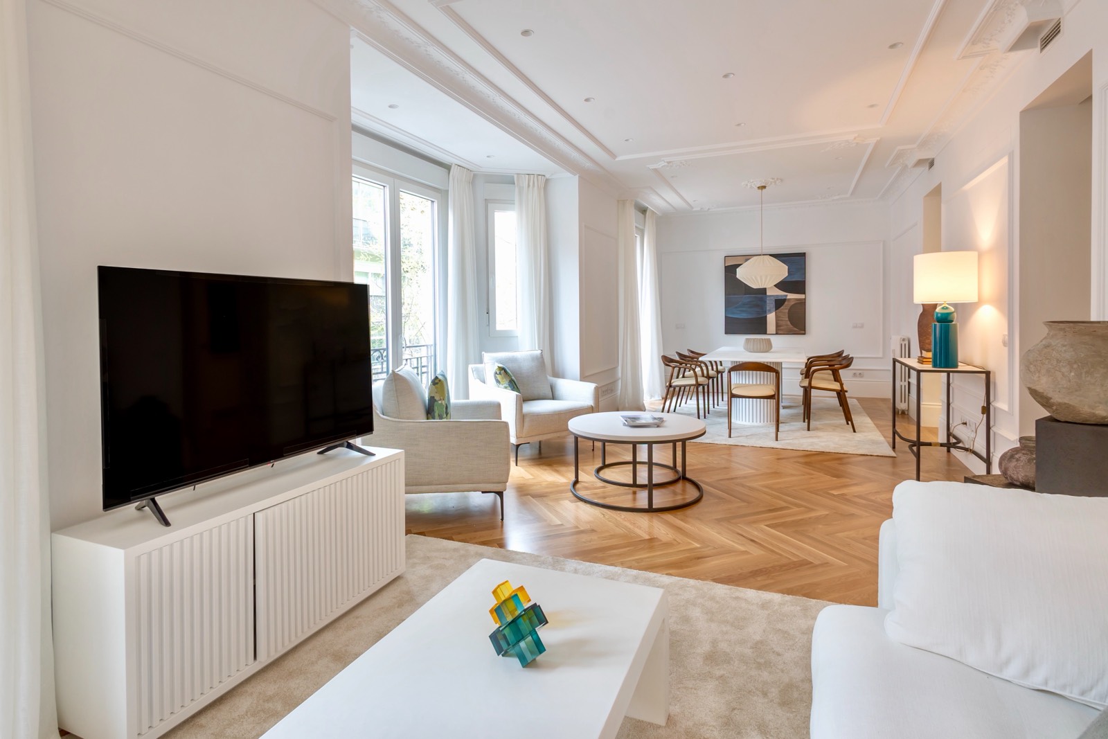 Sala de estar elegante y moderna con decoración en tonos claros, piso de madera en espiga, muebles contemporáneos, televisor grande y ventanales que aportan luz natural.