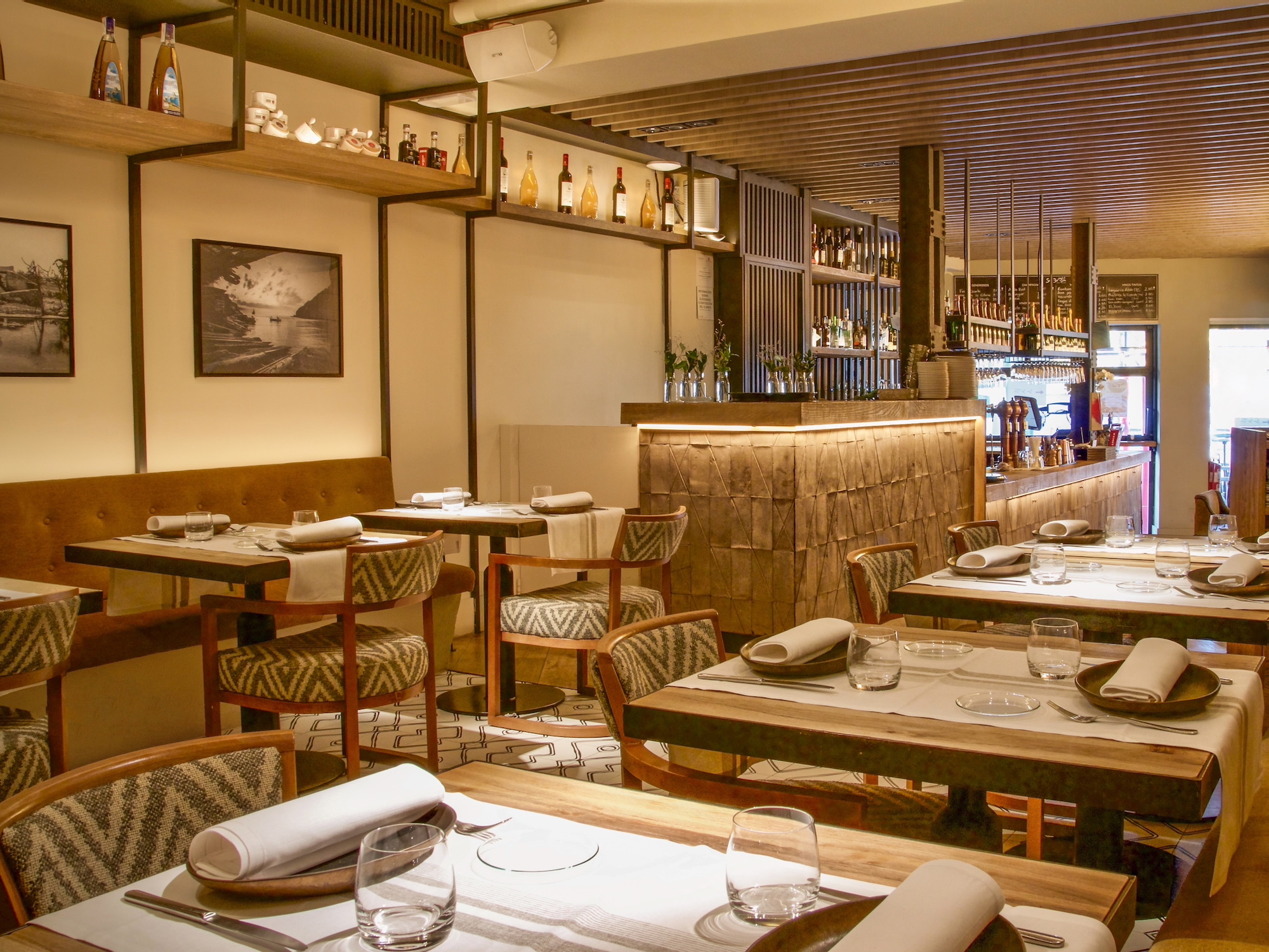 Interior de un restaurante elegante con mesas puestas, sillas tapizadas con patrón geométrico, bar iluminado con estantes de licores, y decoración en tonos cálidos con fotografías en blanco y negro en las paredes