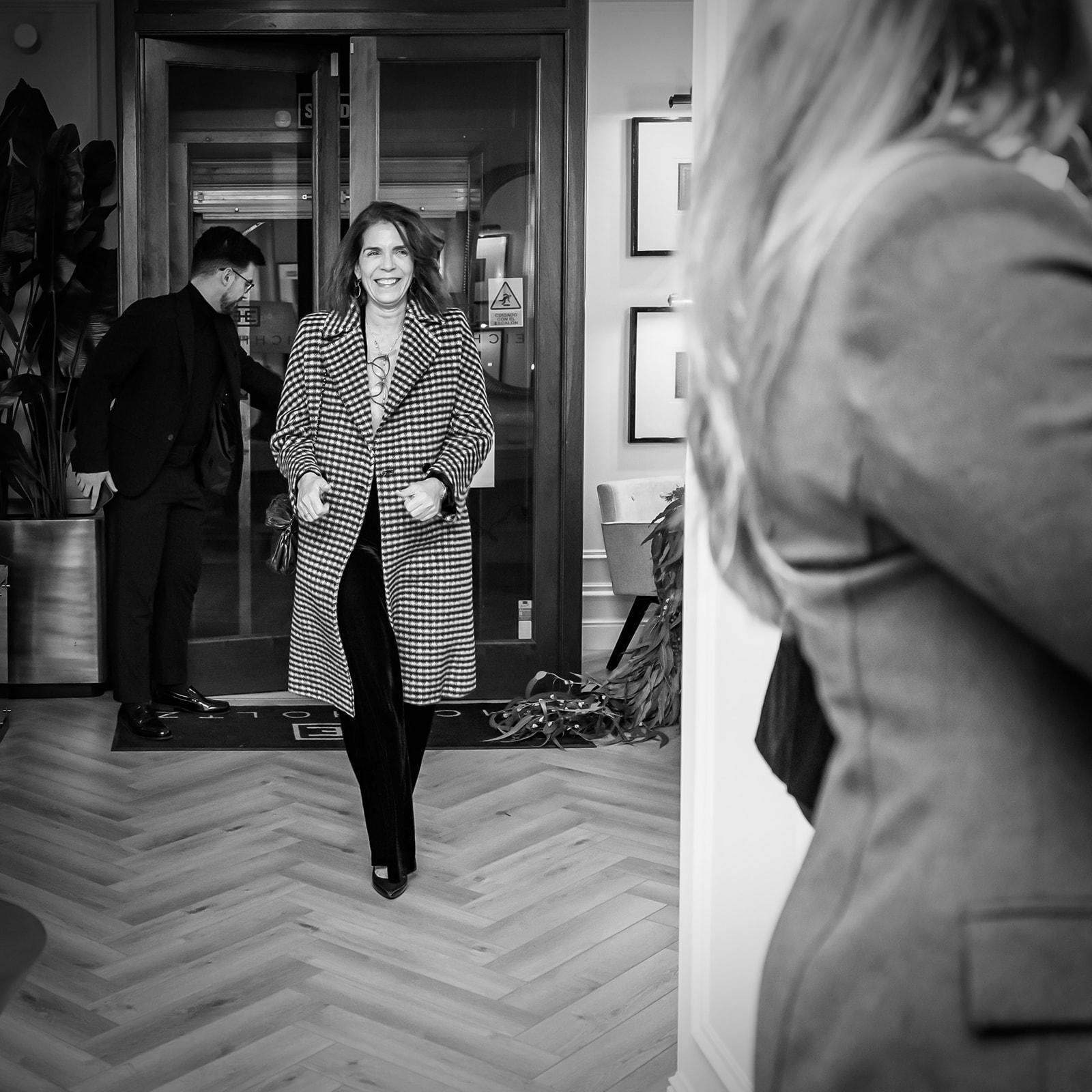 Imagen en blanco y negro de una mujer sonriente entrando por una puerta, vistiendo un abrigo de cuadros. El interior muestra un suelo de parquet y algunas plantas. Otras personas están presentes en el fondo.