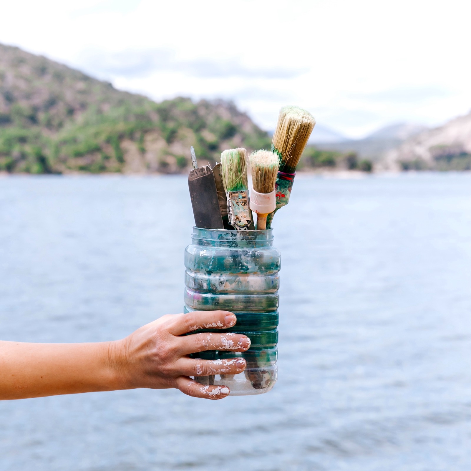 Mano sosteniendo una botella de plástico reutilizada llena de pinceles frente a un hermoso lago rodeado de montañas. Los pinceles están cubiertos de pintura, sugiriendo que se han usado para crear arte al aire libre.