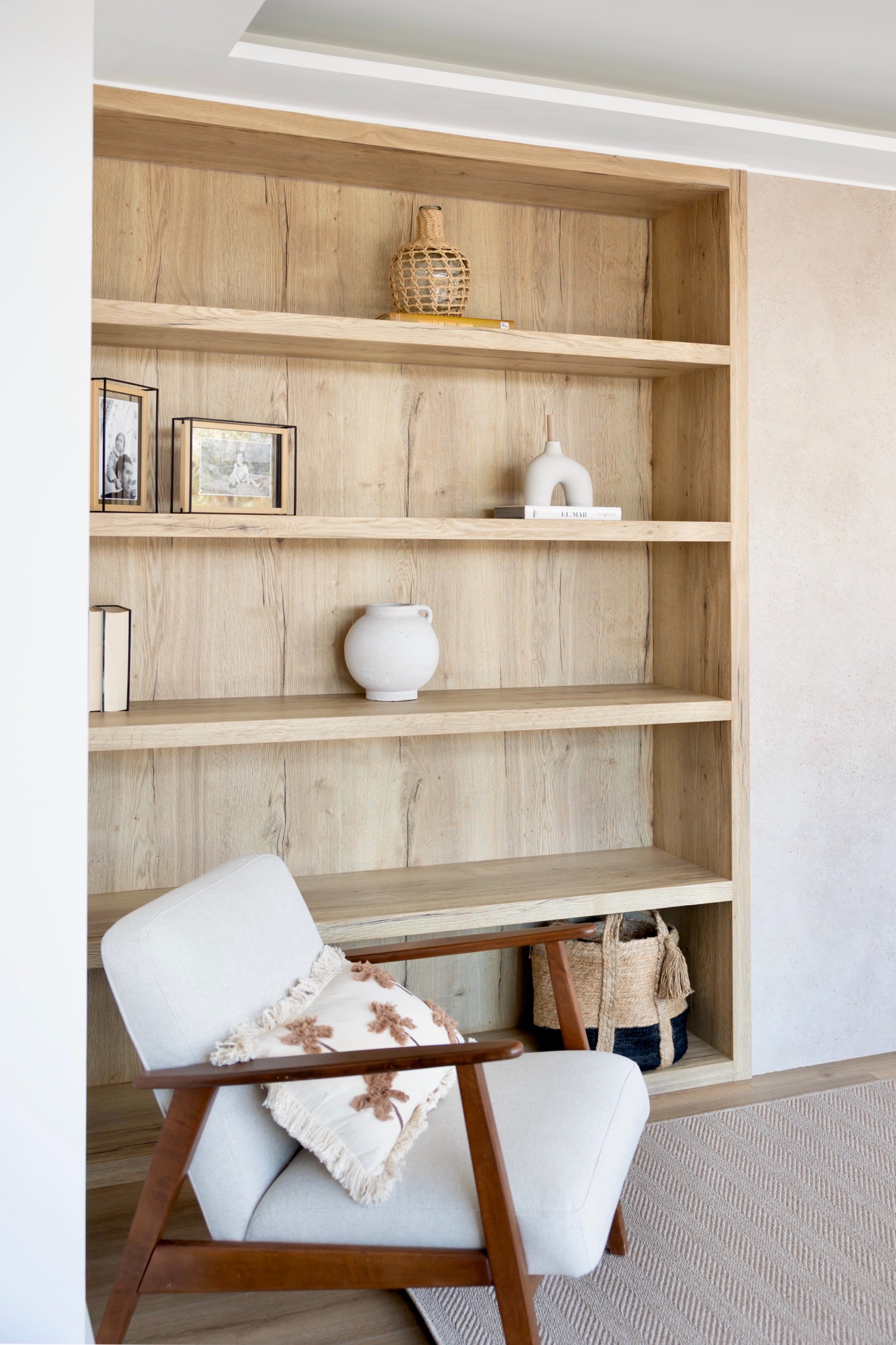 Estantería de madera clara empotrada con varios estantes mostrando decoraciones como jarrones, cuadros y libros. En frente hay un sillón blanco con estructura de madera y un cojín decorativo.