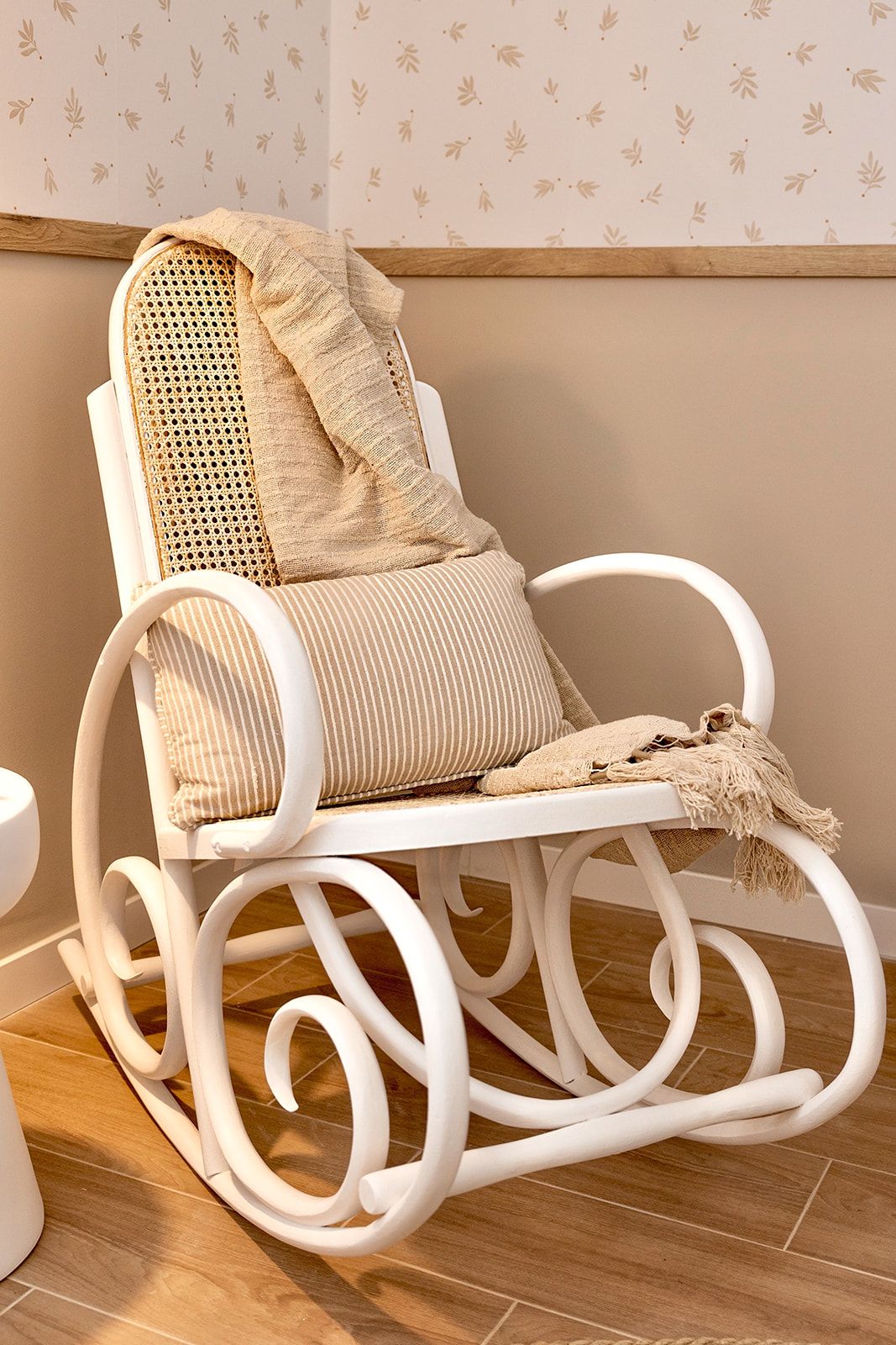 Mecedora blanca de estilo boho con respaldo de rejilla, cojines beige y una manta sobre el asiento. La silla está colocada en una esquina con papel tapiz floral y suelo de madera.