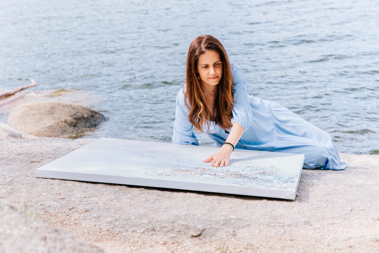 Mujer en vestido azul examina un lienzo blanco con detalles brillantes mientras está sentada en una playa rocosa junto al mar