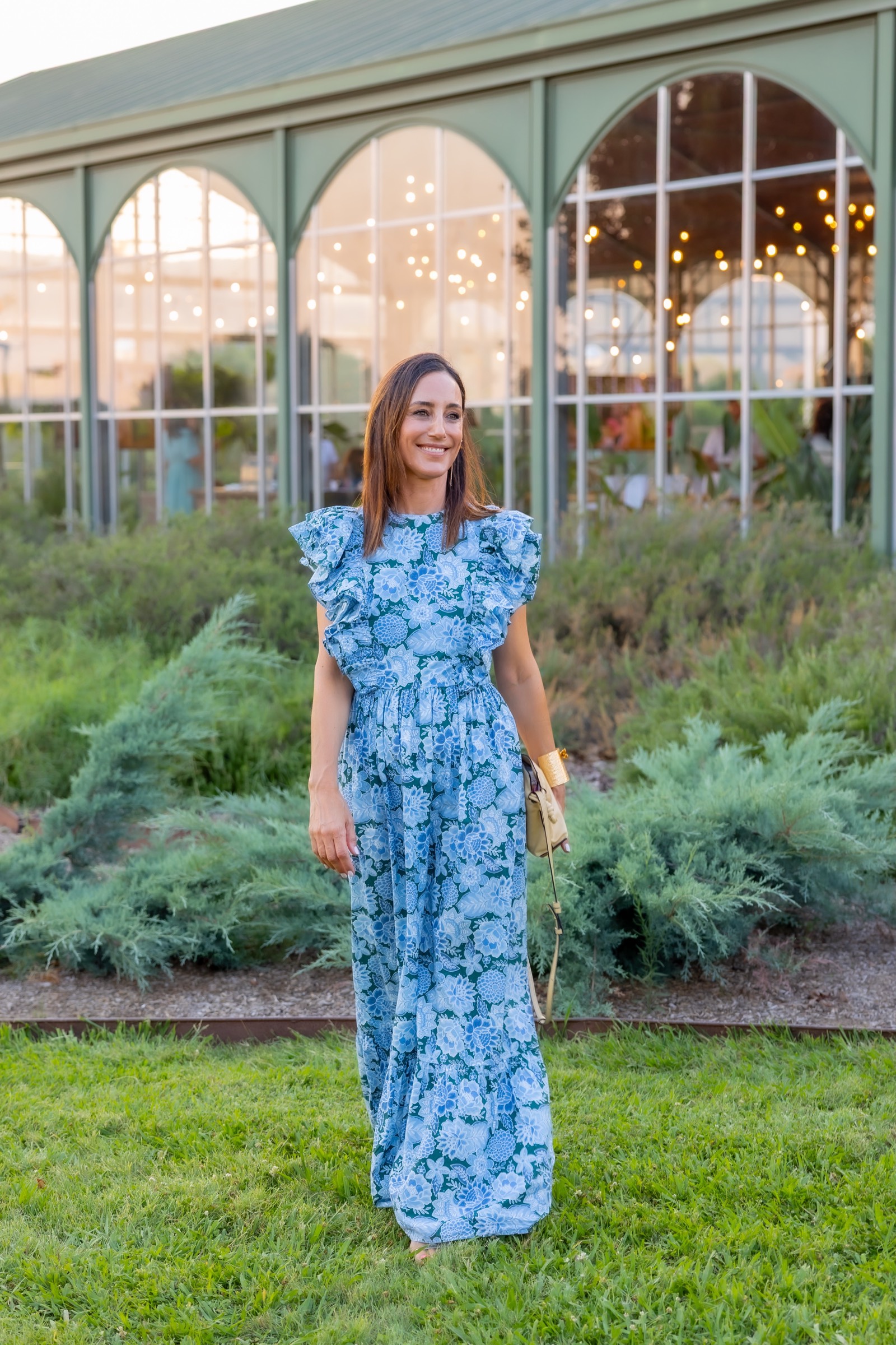 Mujer sonriente con vestido largo floral azul, de pie en un jardín con edificio de invernadero con luces al fondo