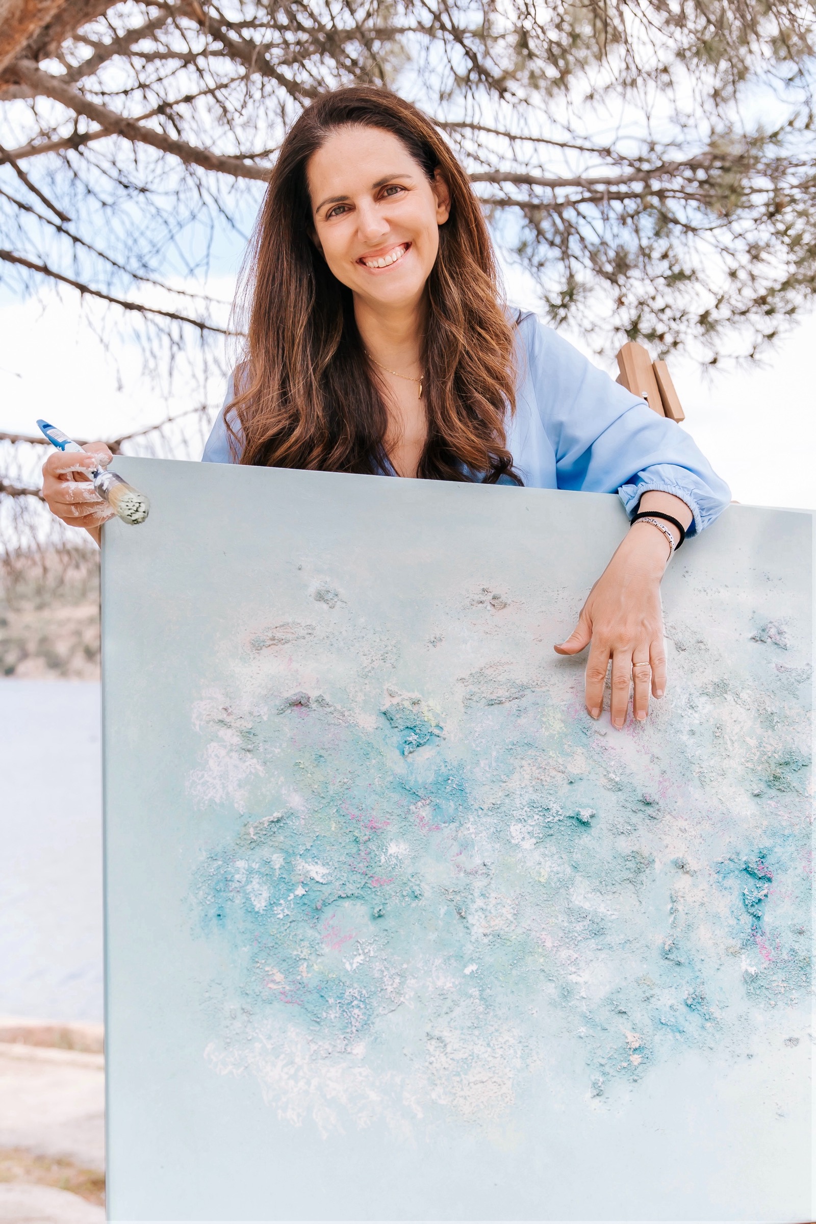 Mujer sonriente sosteniendo una pintura abstracta en tonos pastel azules y rosas. Está al aire libre bajo un árbol en un día soleado, vistiendo una camisa azul claro.