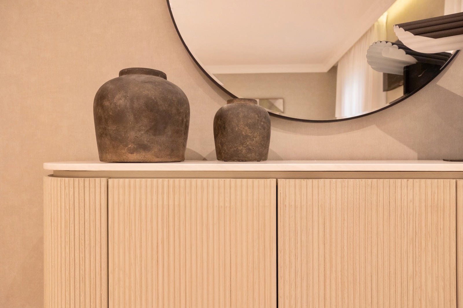 Dos jarrones rústicos de color marrón oscuro colocados sobre un mueble de madera clara con puertas acanaladas. Un gran espejo redondo cuelga en la pared sobre el mueble, reflejando parte de la habitación.