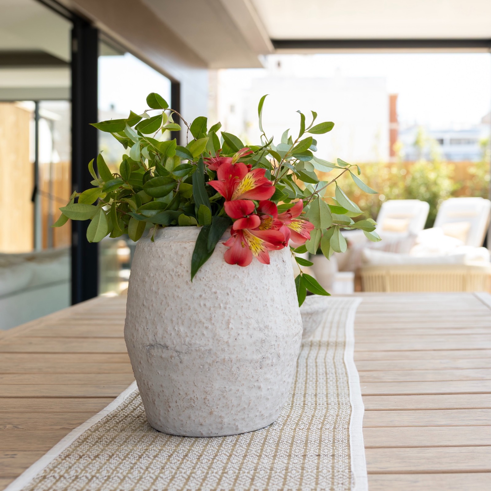 Arreglo floral con flores rojas y hojas verdes en un jarrón blanco sobre una mesa de madera en una terraza al aire libre con muebles de jardín al fondo