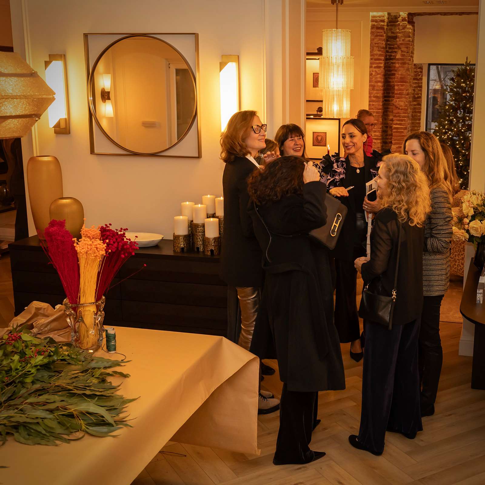 Grupo de personas conversando animadamente en un salón decorado para las fiestas, con velas, flores y un espejo circular. El ambiente es cálido y acogedor, con iluminación tenue y decoración navideña.
