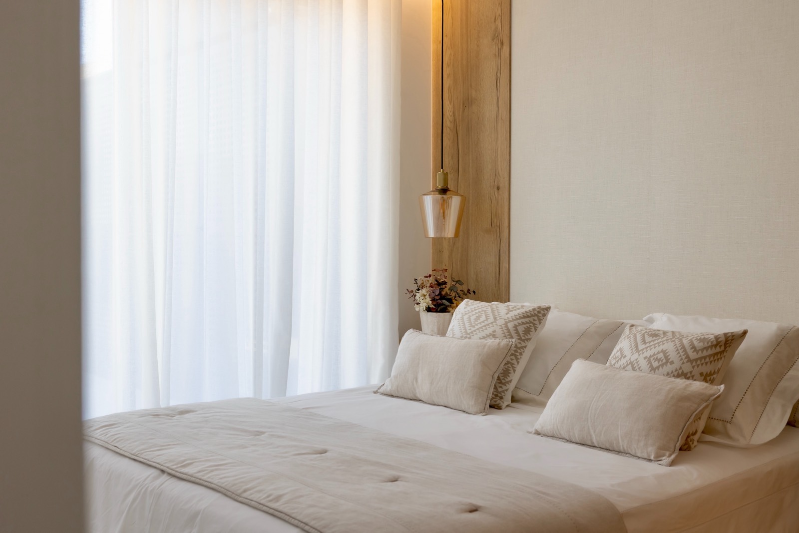 Dormitorio luminoso con estilo escandinavo, cama blanca con cojines decorativos, lámpara colgante de cristal y madera, cortinas blancas translúcidas y un pequeño jarrón con flores.