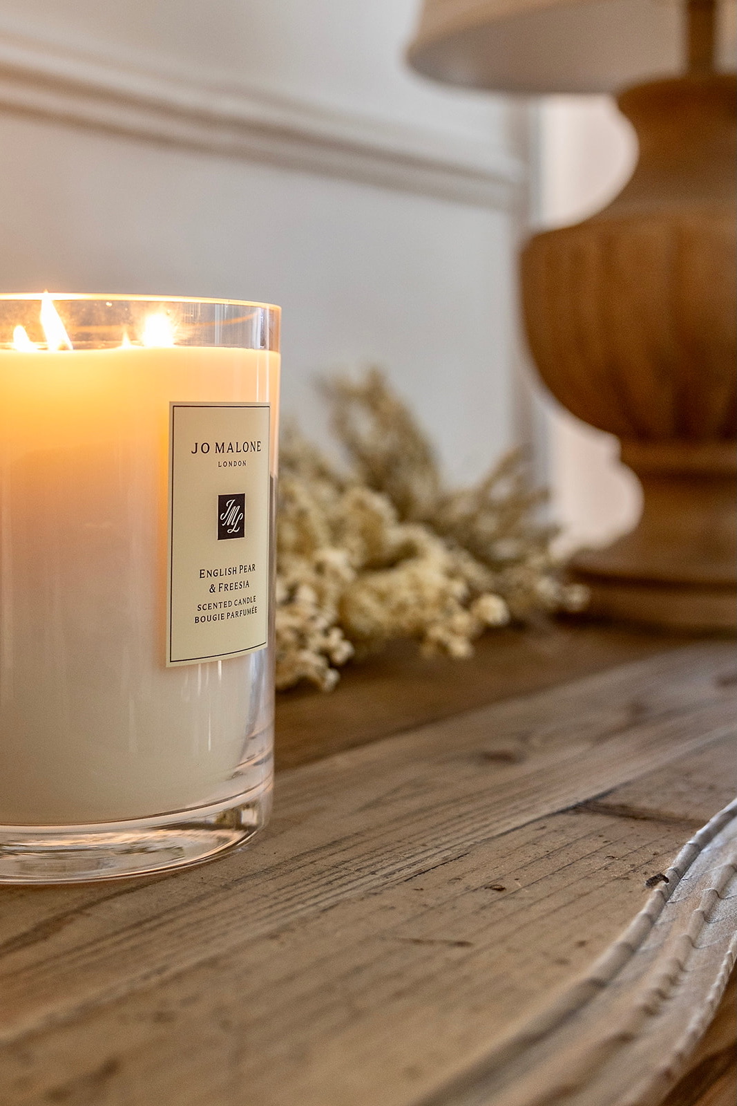Vela aromática Jo Malone de English Pear & Freesia encendida sobre una superficie de madera rústica con flores secas decorativas en el fondo, creando una atmósfera acogedora y elegante