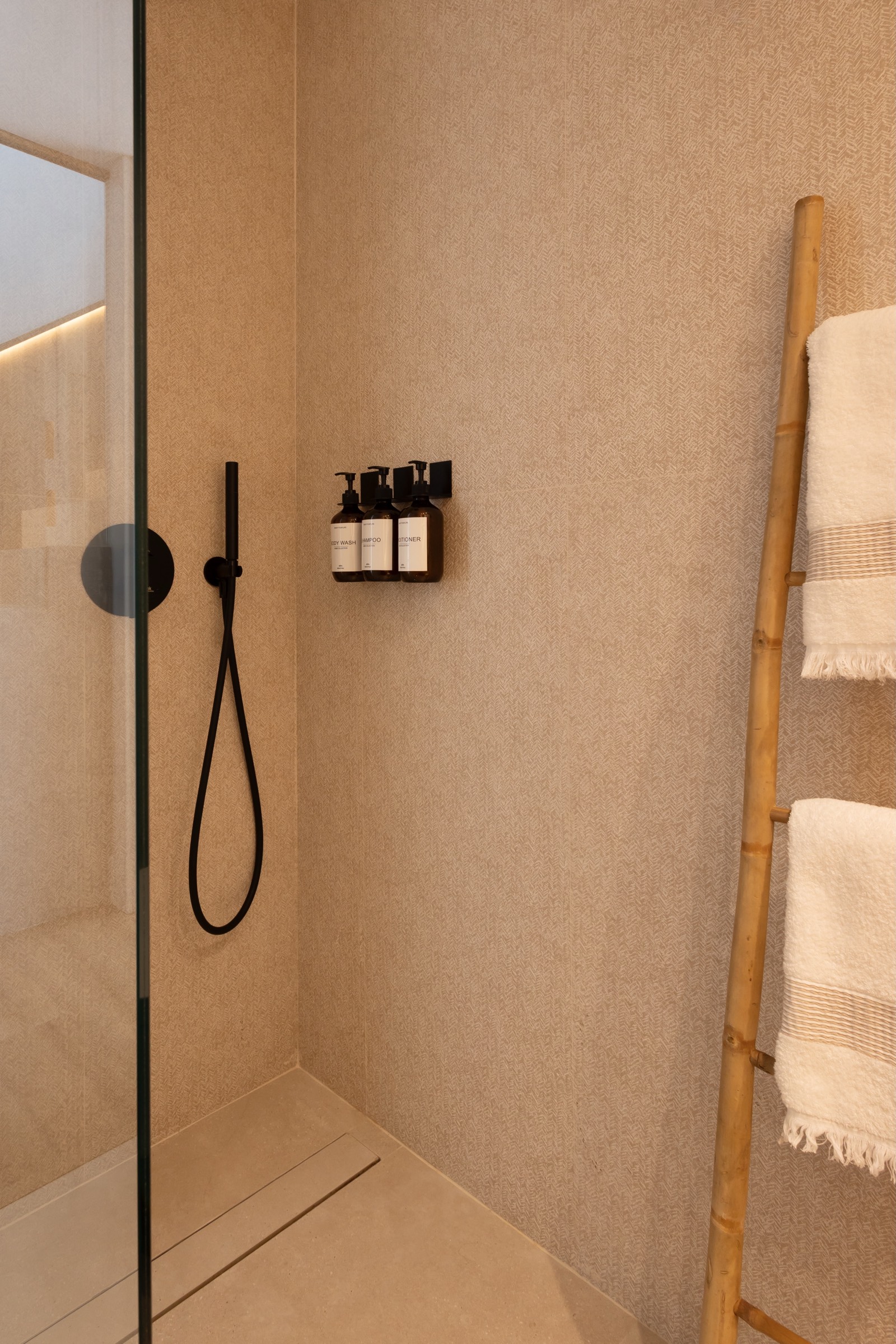 Ducha moderna y minimalista con paredes de color beige, accesorios negros, estante con botellas de productos y una escalera de bambú con toallas blancas.