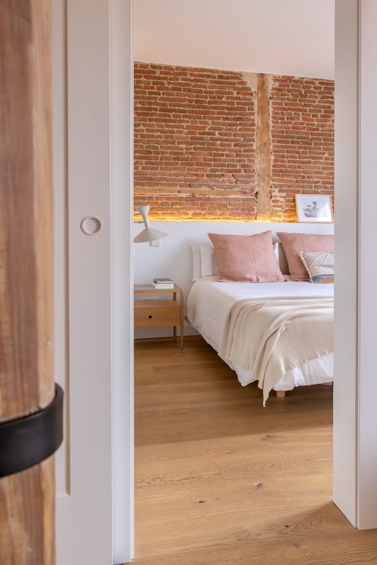 Interior de dormitorio moderno con pared de ladrillo expuesto, cama blanca con cojines rosados y suelo de madera clara