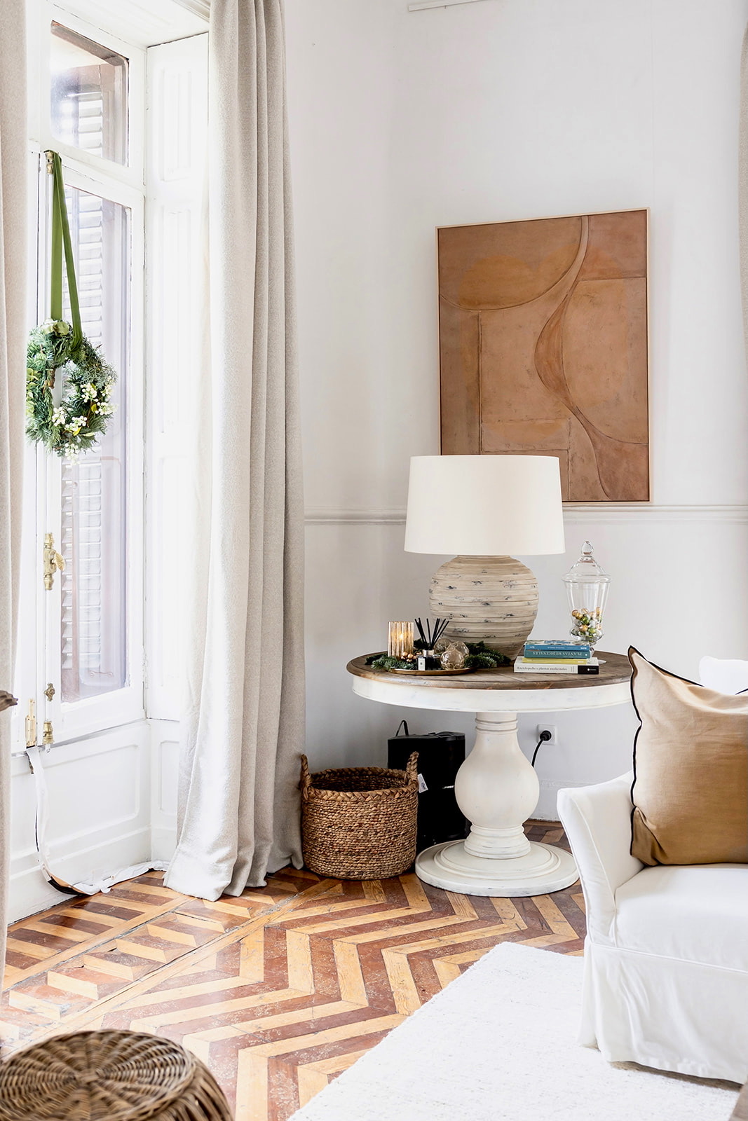 Sala de estar luminosa con pisos de madera en patrón chevron, mesa lateral redonda blanca, lámpara cerámica, corona navideña en ventana y arte abstracto en tonos tierra. Decoración en estilo costero con elementos naturales.