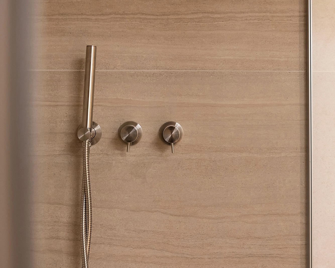 Ducha moderna con grifería de acero inoxidable montada en la pared. Incluye una alcachofa de mano y dos mandos circulares sobre un fondo de azulejos beige con textura de piedra.