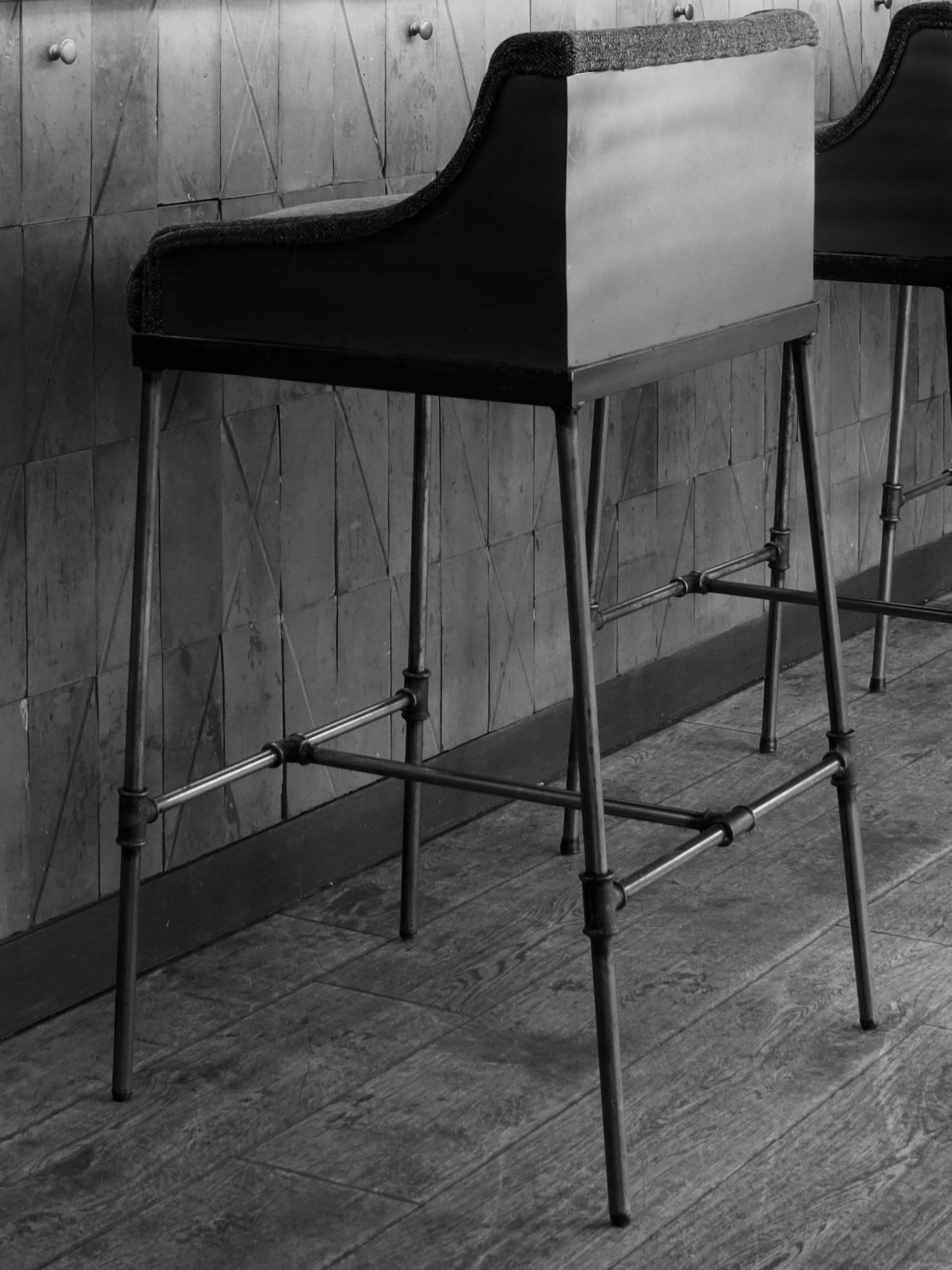 Taburete alto de bar en estilo industrial con respaldo negro, estructura metálica y patas cruzadas, fotografiado en blanco y negro contra una pared texturizada