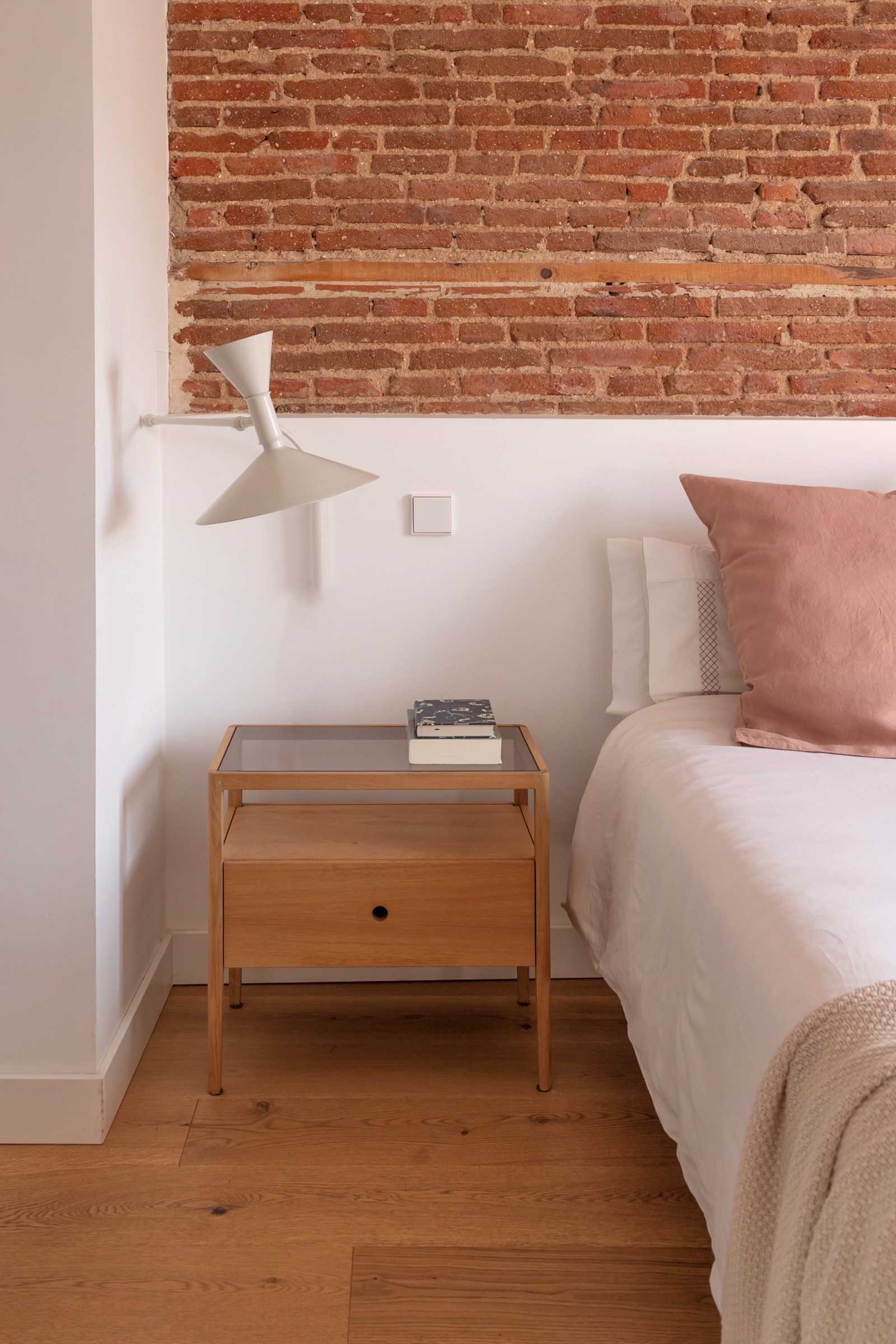 Rincón de dormitorio minimalista con pared de ladrillo expuesto, mesita de noche de madera, lámpara blanca y cama con cojines en tonos rosados
