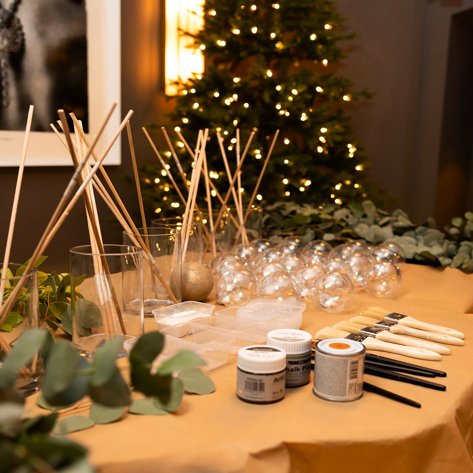 Mesa preparada para un taller de manualidades navideñas con materiales como bolas de cristal, palos de madera, pinturas, pinceles y ramas de eucalipto. Al fondo se ve un árbol de Navidad iluminado.