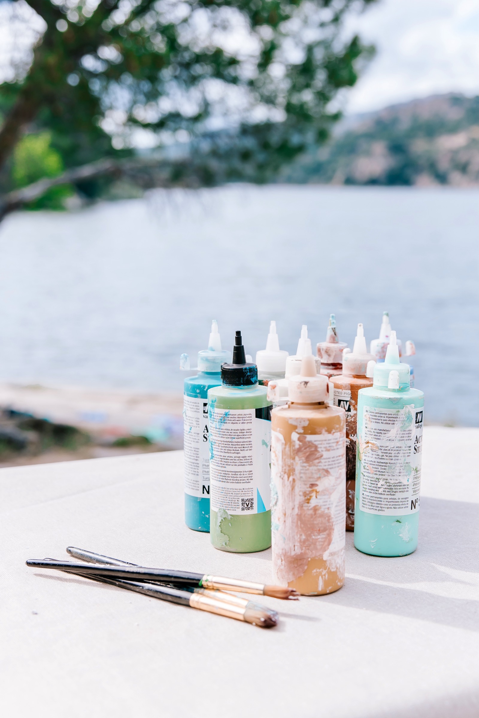 Botellas de pintura acrílica y pinceles sobre una mesa blanca junto a un lago tranquilo. Al fondo se ven árboles y montañas borrosas. Las pinturas son de colores pastel como turquesa, verde claro y rosa.