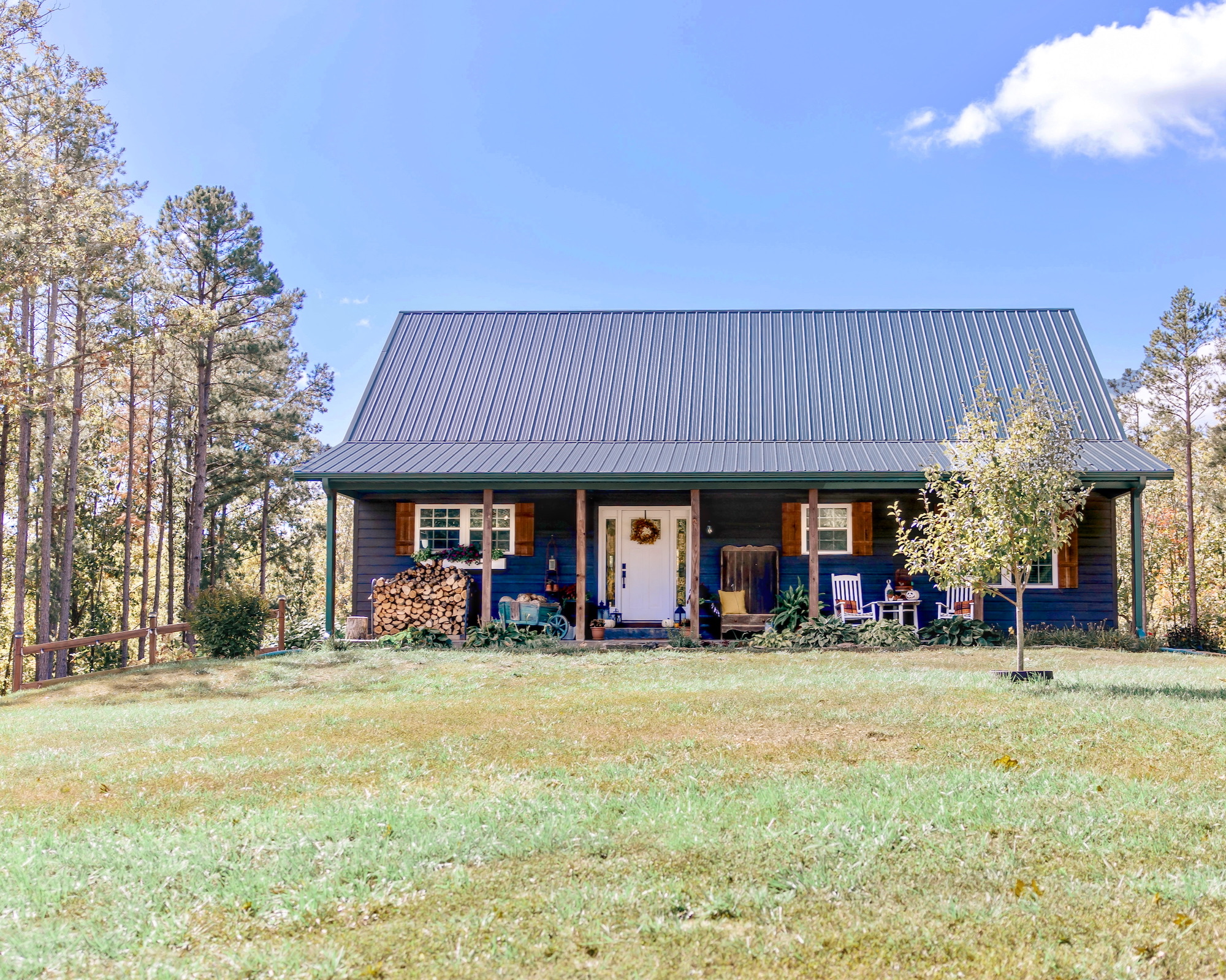 Casa de estilo rústico pintada de azul con techo de metal gris, porche frontal decorado con mecedoras y leña apilada, rodeada de pinos y césped natural en un entorno boscoso