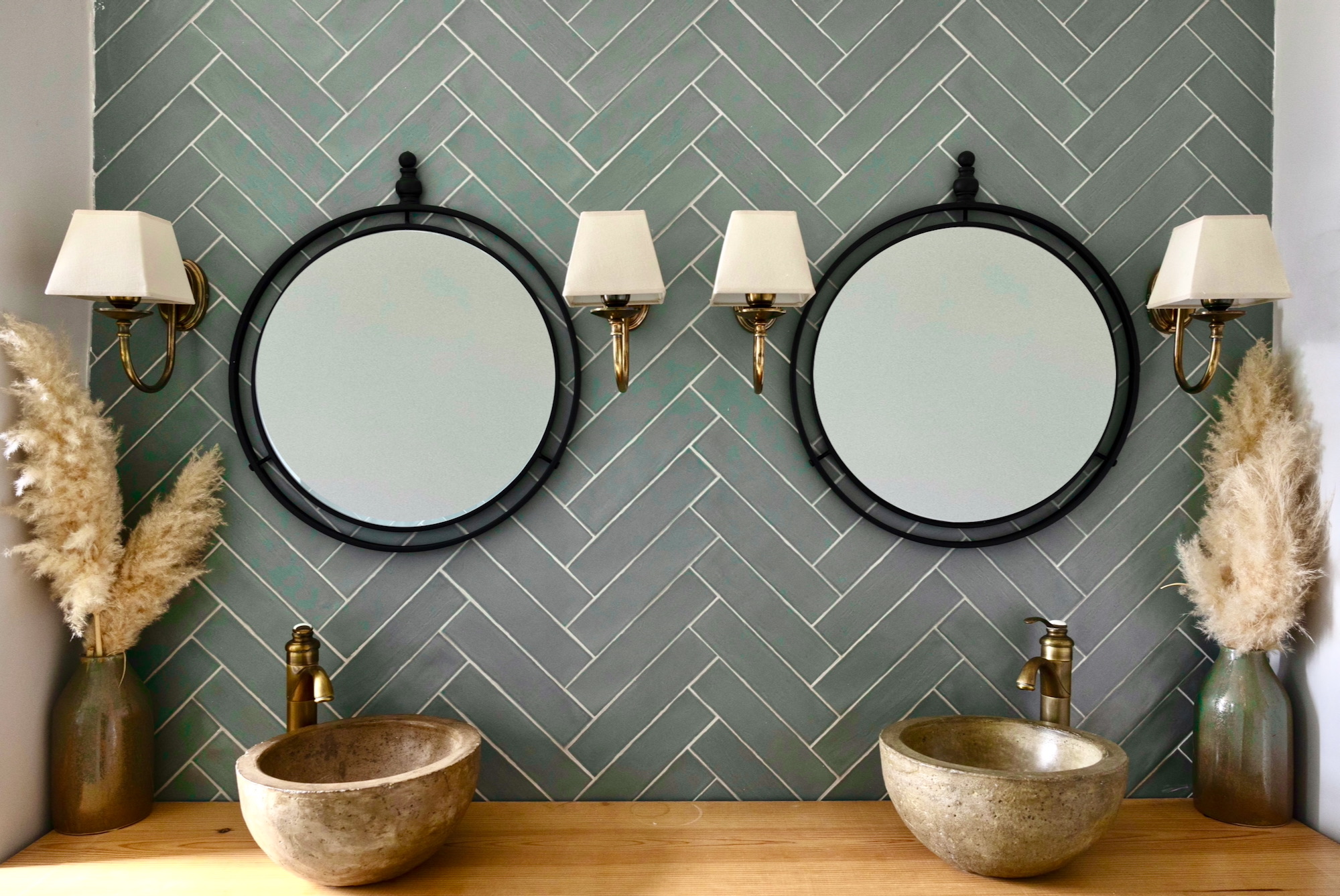 Baño moderno con azulejos verdes en patrón espiga, dos espejos redondos negros, lavabos tipo bowl de concreto sobre encimera de madera, apliques dorados y jarrones decorativos con pampas