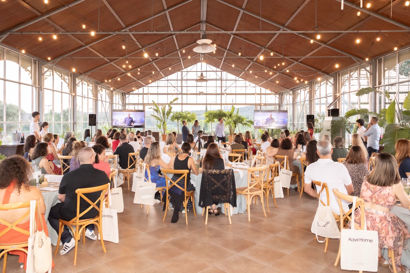 Sala de evento con asistentes sentados en sillas de madera, iluminación con luces colgantes y pantallas en el escenario