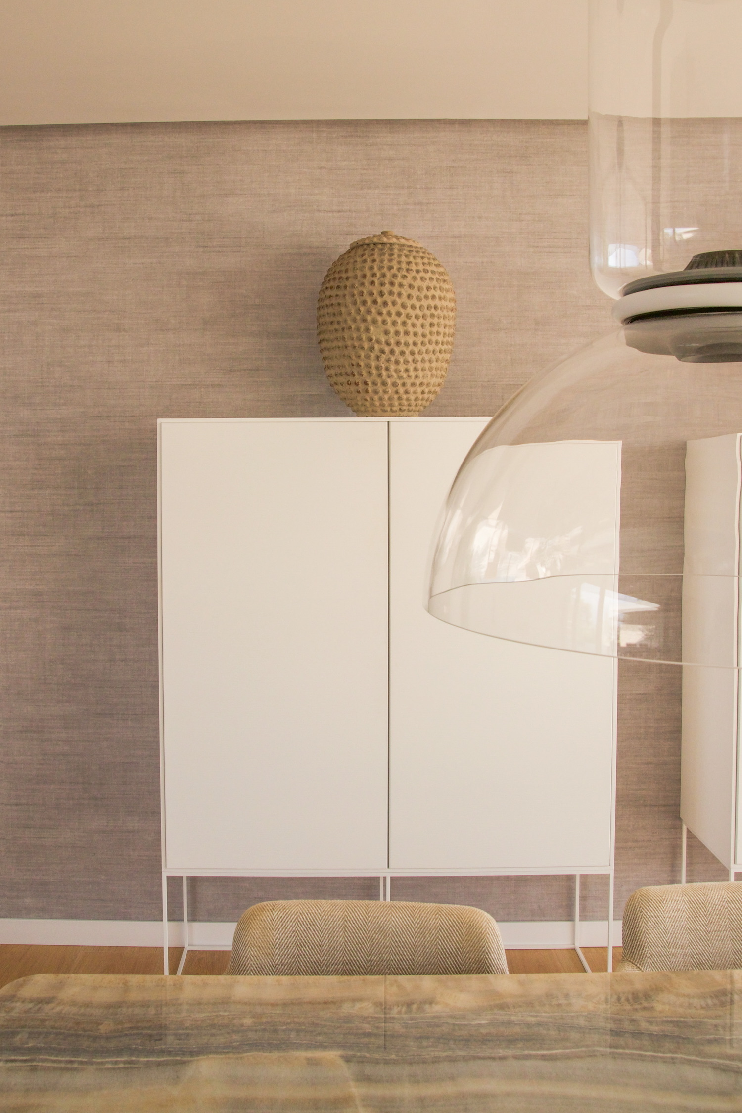 Diseño interior minimalista de comedor con gabinete blanco, jarrón decorativo texturizado, lámpara colgante de cristal y mesa de madera con silla tapizada en tono beige