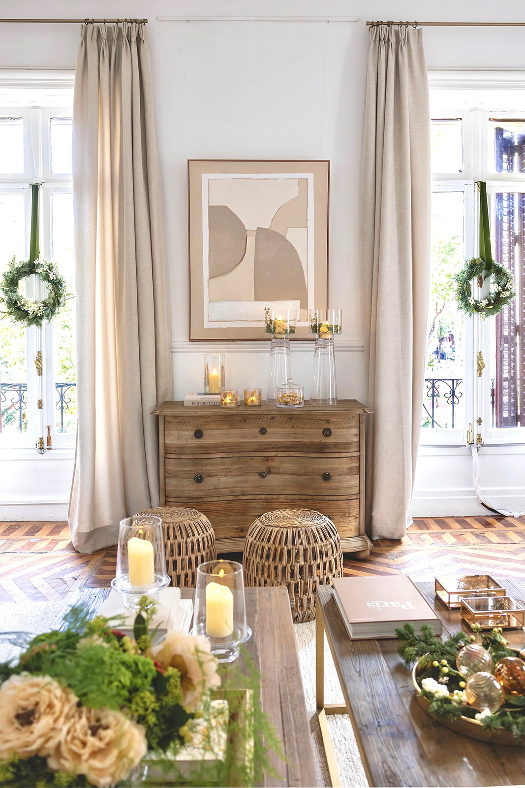Sala de estar con decoración navideña elegante en tonos neutros, con cómoda de madera rústica, velas, coronas verdes en las ventanas, taburetes de ratán y cortinas beige. Arte abstracto en la pared y detalles decorativos festivos.