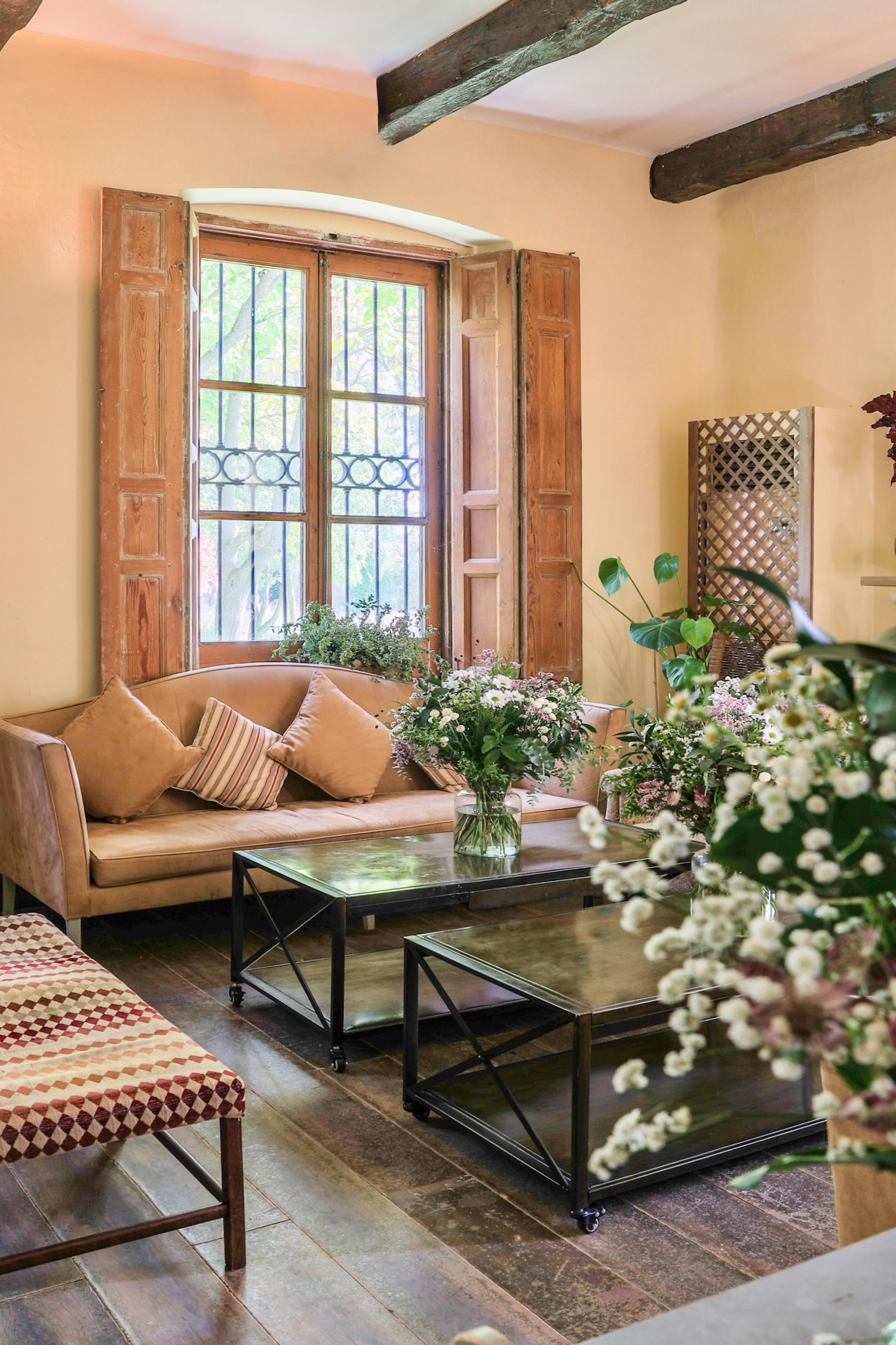 Sala de estar rústica mediterránea con ventana alta y contraventanas de madera, sofá beige con cojines, mesas de centro negras y decoración floral. Vigas de madera en el techo y suelo de terracota.