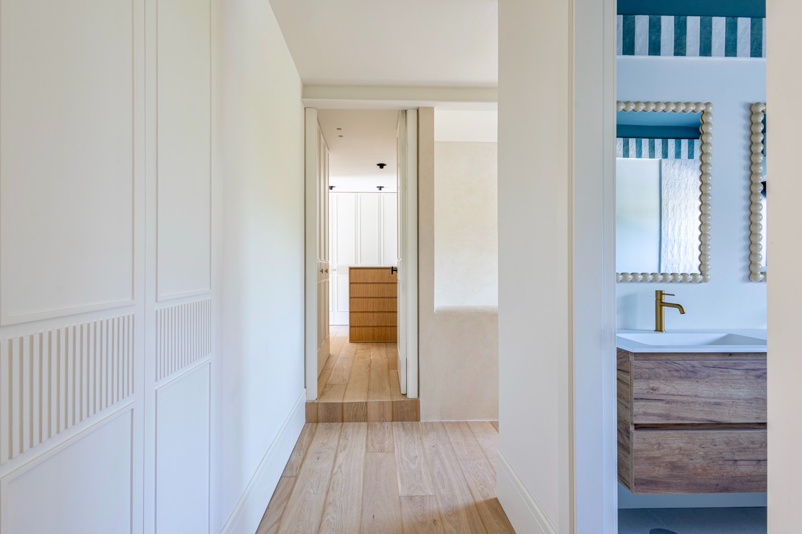 Interior de un pasillo luminoso con paredes blancas, suelo de madera clara y vista parcial de un baño con lavabo y espejo