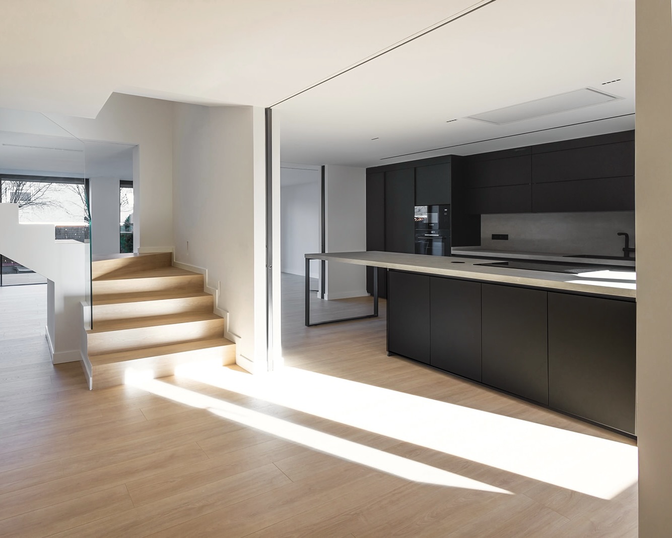 Interior de una casa moderna con una cocina minimalista de color negro y blanco, y escaleras iluminadas que conducen a otro nivel. El espacio está bañado en luz natural, creando un ambiente luminoso y acogedor.