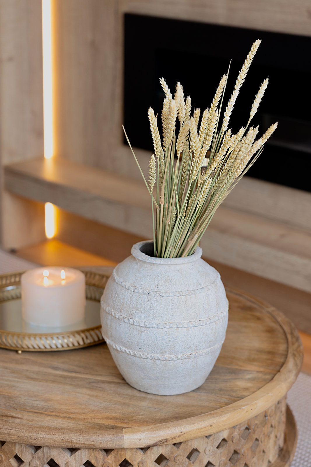 Florero blanco con espigas de trigo seco sobre una mesa de madera redonda. Una vela encendida junto al florero. Ambiente acogedor con iluminación cálida.