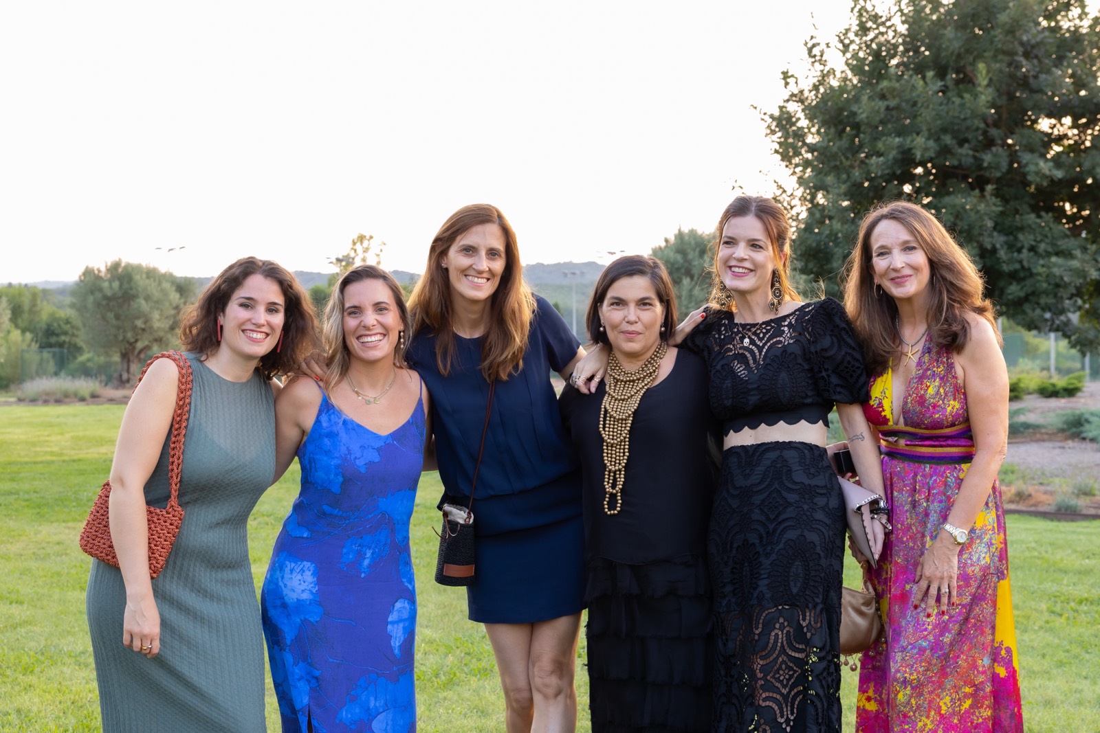 Seis mujeres sonrientes vestidas con diferentes estilos de vestidos, posando juntas en un jardín con árboles y césped al fondo
