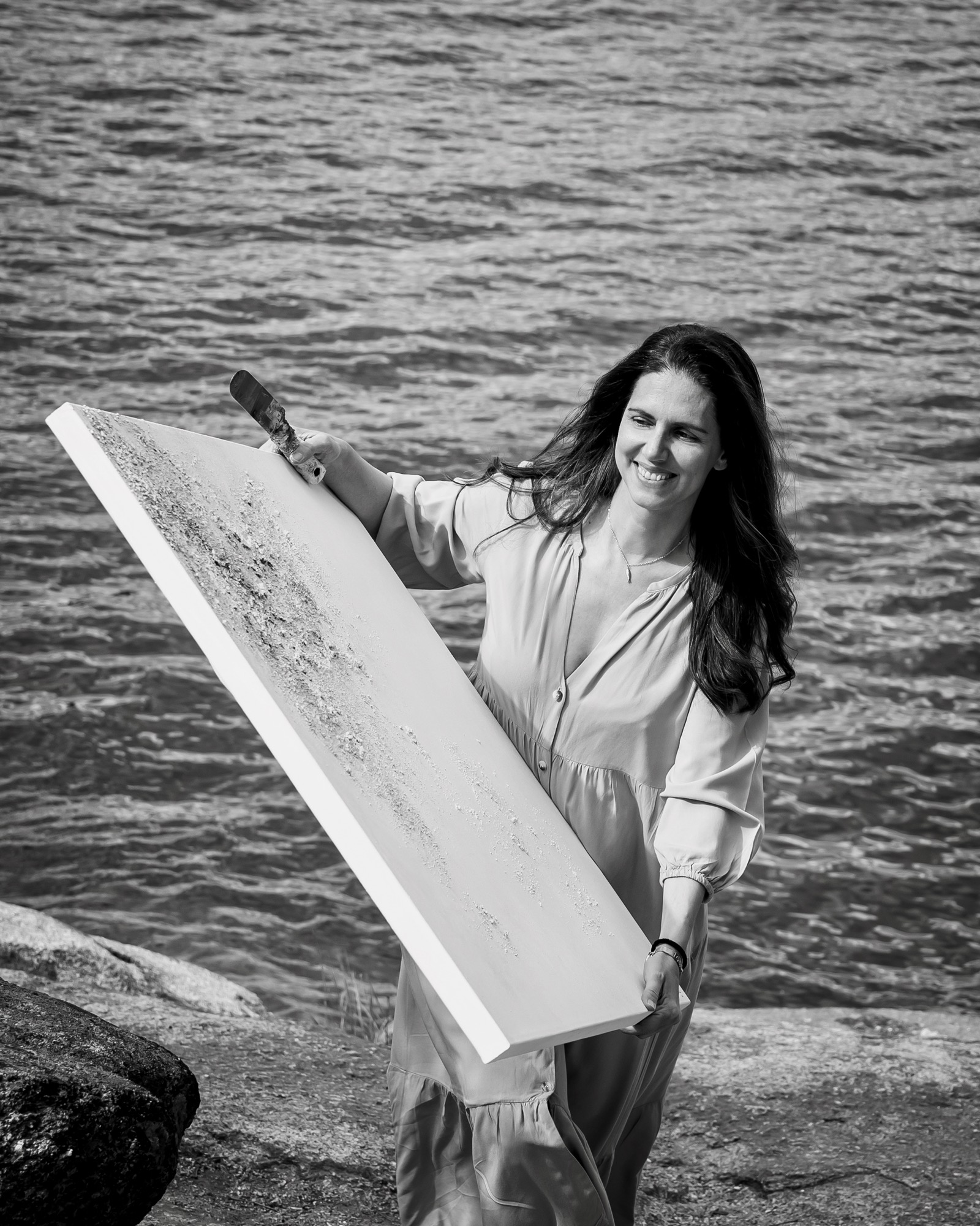 Artista sonriente sosteniendo un gran lienzo blanco con pintura y pincel junto al mar en una imagen en blanco y negro. La mujer lleva un vestido largo y su cabello ondea con la brisa marina.