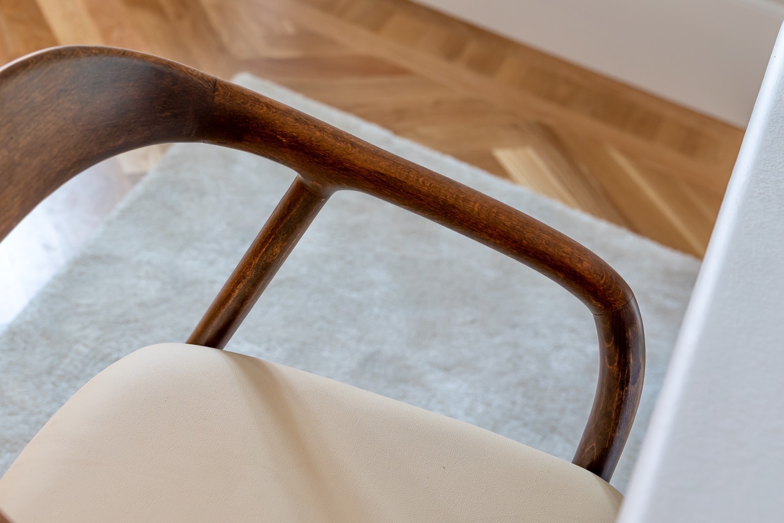 Detalle de una silla de diseño escandinavo con reposabrazos de madera curvada y asiento tapizado en cuero beige. Se aprecia el parqué de madera en el fondo y una alfombra gris.