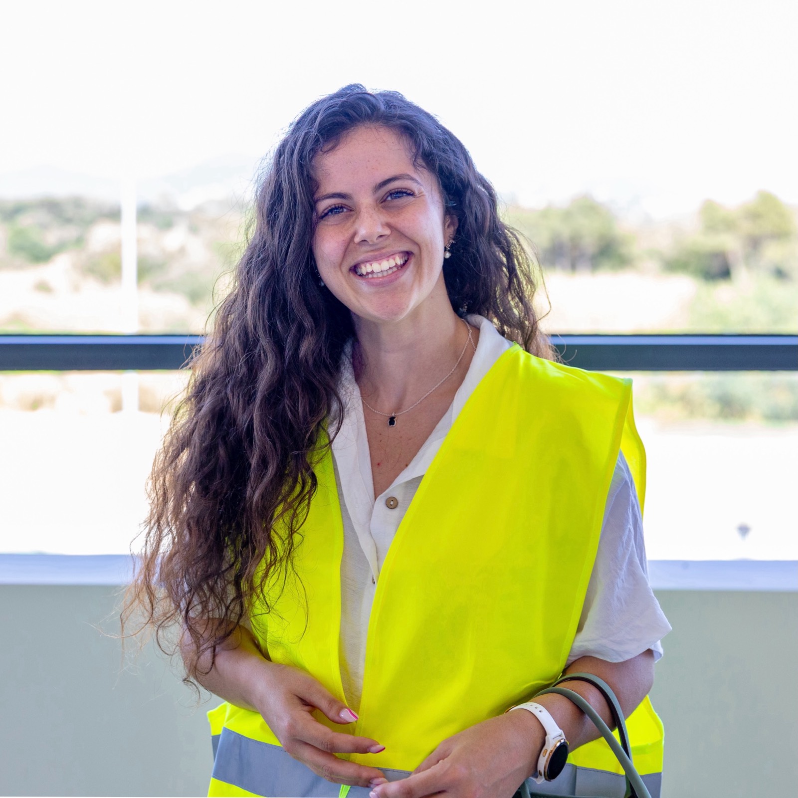 Mujer joven con chaleco de seguridad amarillo brillante, sonriendo ampliamente con cabello rizado largo, en un entorno con vista al exterior