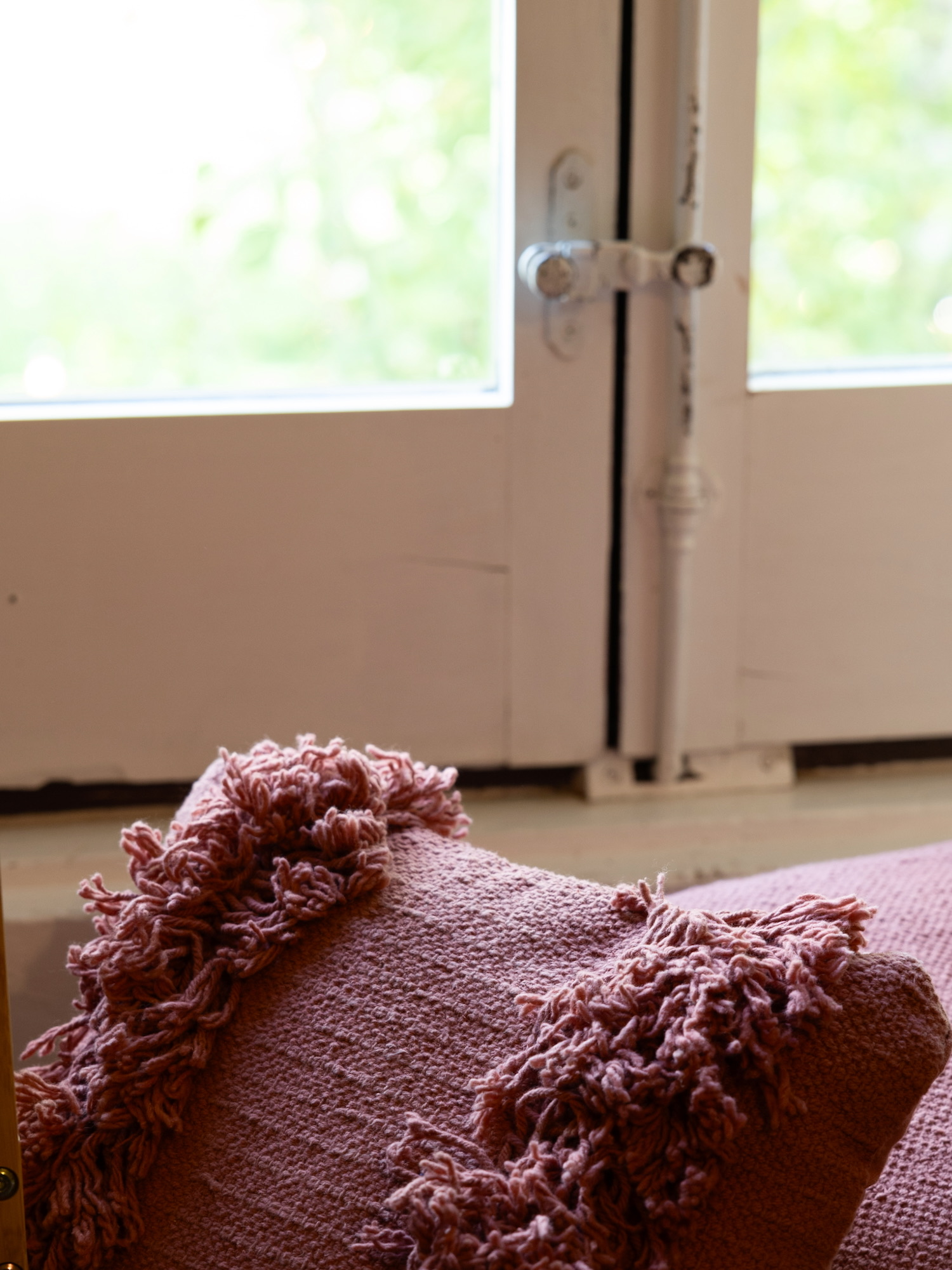 Cojín decorativo de color rosa con flecos ornamentales en los bordes, colocado junto a una ventana con luz natural filtrada, creando una atmósfera acogedora y hogareña