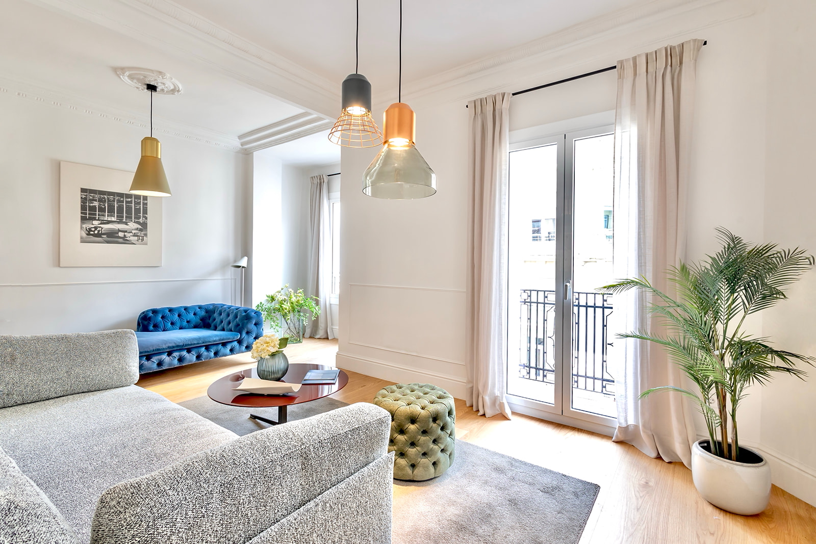 Sala de estar moderna y elegante con sofá azul, lámparas colgantes de colores, ventanas grandes con cortinas claras, y elementos decorativos como plantas y un puf verde. El espacio es luminoso y acogedor.