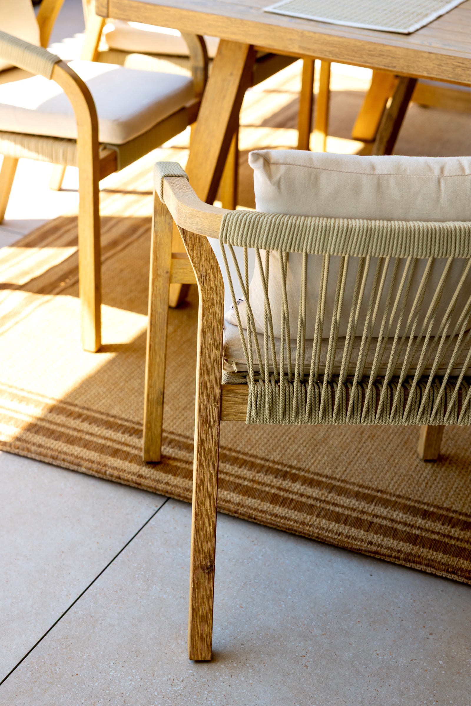 Detalle de muebles de terraza de madera con sillas de cuerda. Se muestra una mesa y sillas de madera clara, con una silla de cuerda en primer plano. Hay una alfombra beige debajo y el ambiente está bañado en luz solar.