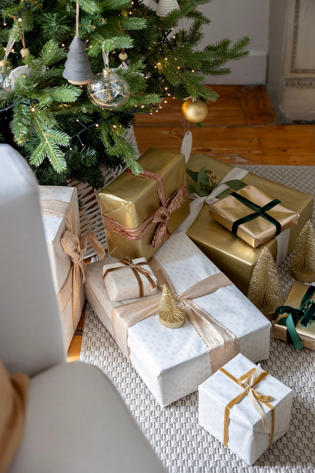 Regalos navideños envueltos en papel dorado y blanco con cintas decorativas, colocados bajo un árbol de Navidad decorado con adornos y luces. Los paquetes están dispuestos sobre una alfombra texturizada.