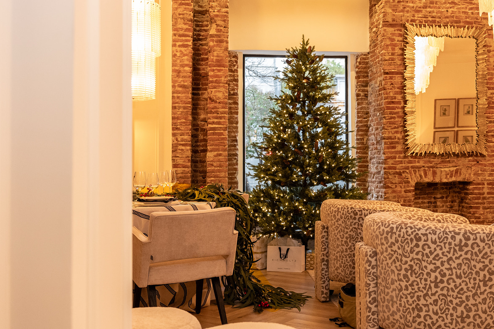 Sala de estar con decoración navideña y estilo rústico. Árbol de Navidad iluminado frente a una ventana, paredes de ladrillo expuesto, sillones estampados y mesa de comedor. Ambiente cálido y acogedor.