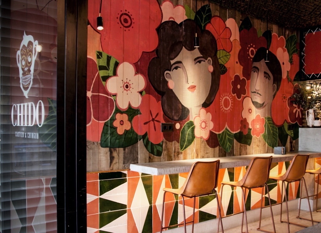 Interior de restaurante mexicano con mural artístico en tonos coral y verde sobre pared de madera, mostrando ilustraciones florales y rostros estilizados. Barra con sillas naranjas y diseño geométrico en la base