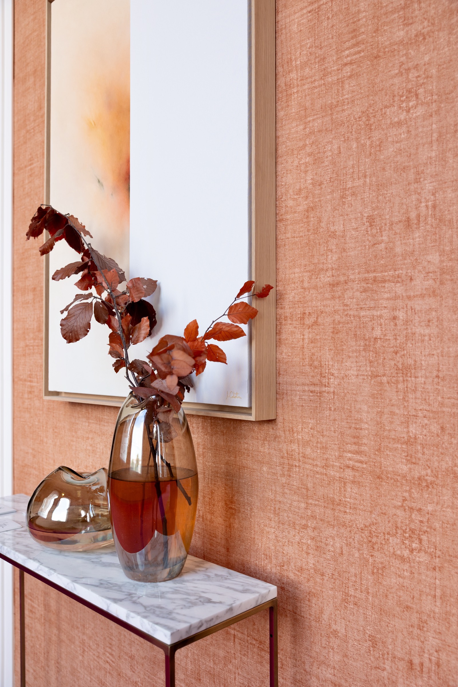 Jarrón de vidrio transparente con ramas de hojas marrones y rojas sobre una mesa de mármol blanco con pared terracota de fondo