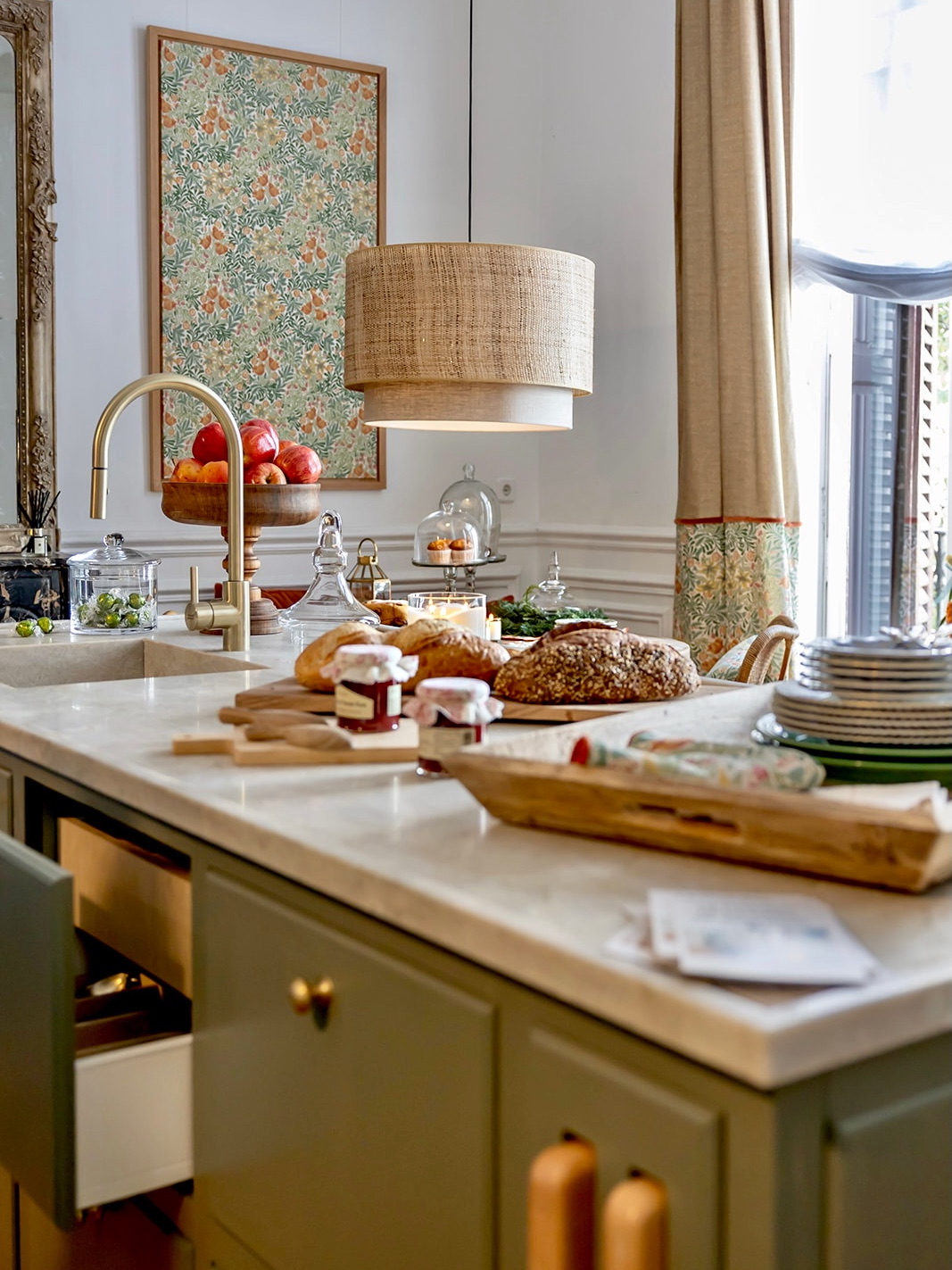Cocina elegante con gabinetes verde oliva, encimera de mármol blanco, lámpara colgante de ratán y cuadro floral vintage. Mesa con panes artesanales, mermeladas y accesorios de cocina dorados