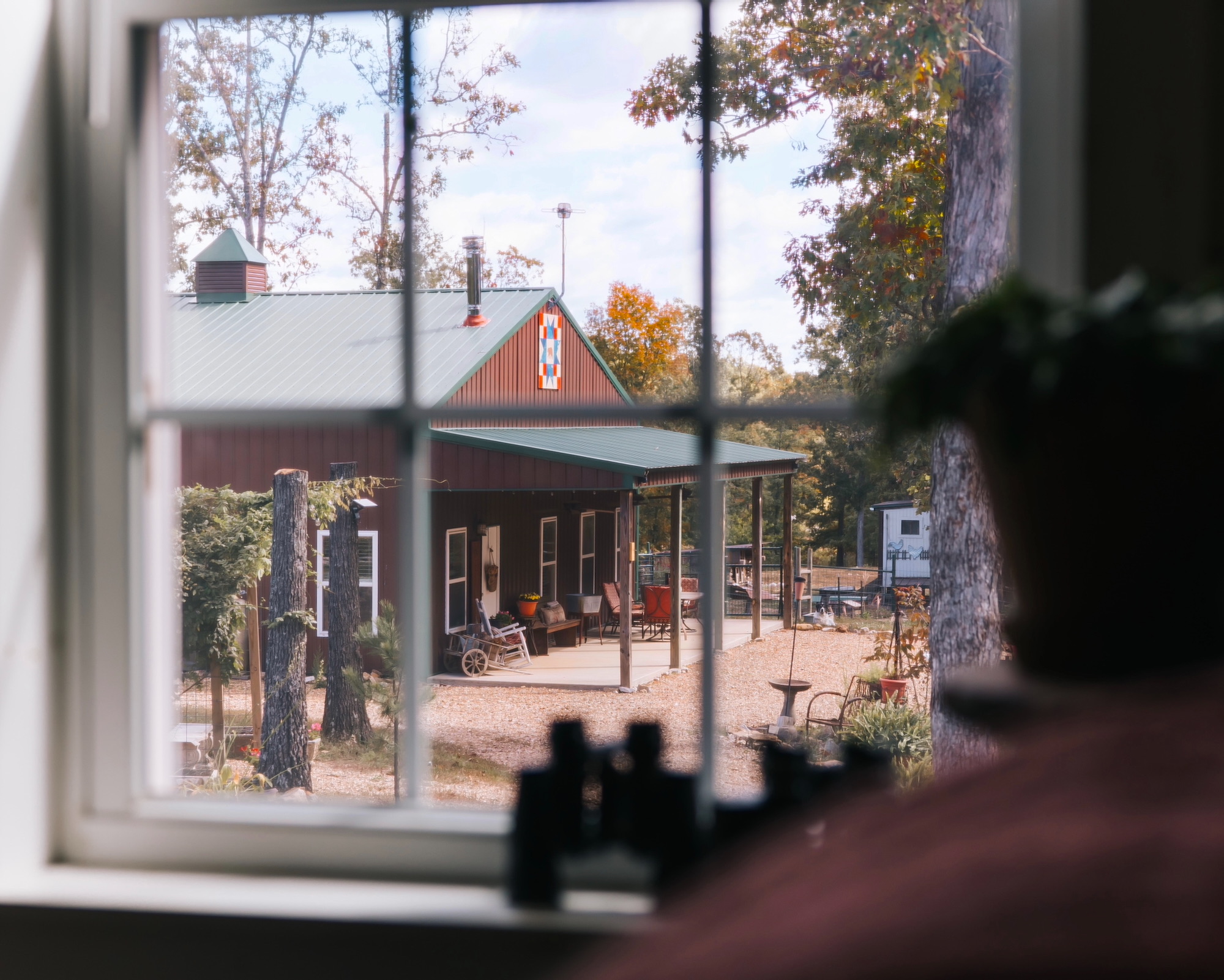 Vista a través de una ventana de una cabaña rústica con porche cubierto, techo metálico verde y paredes marrones. El porche está decorado con muebles de exterior y rodeado de árboles otoñales.